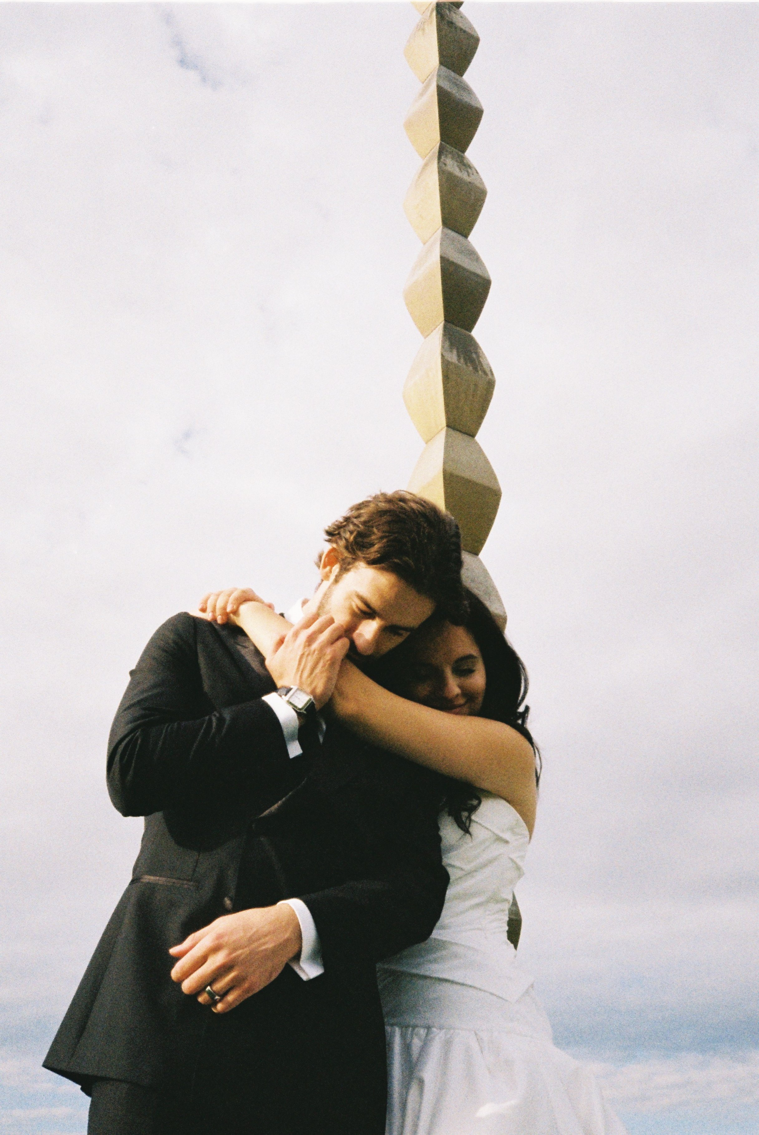 A man and woman embrace and smile passionately outdoors, with the man in a suit and the woman in a white dress, and a tall abstract sculpture in the background.