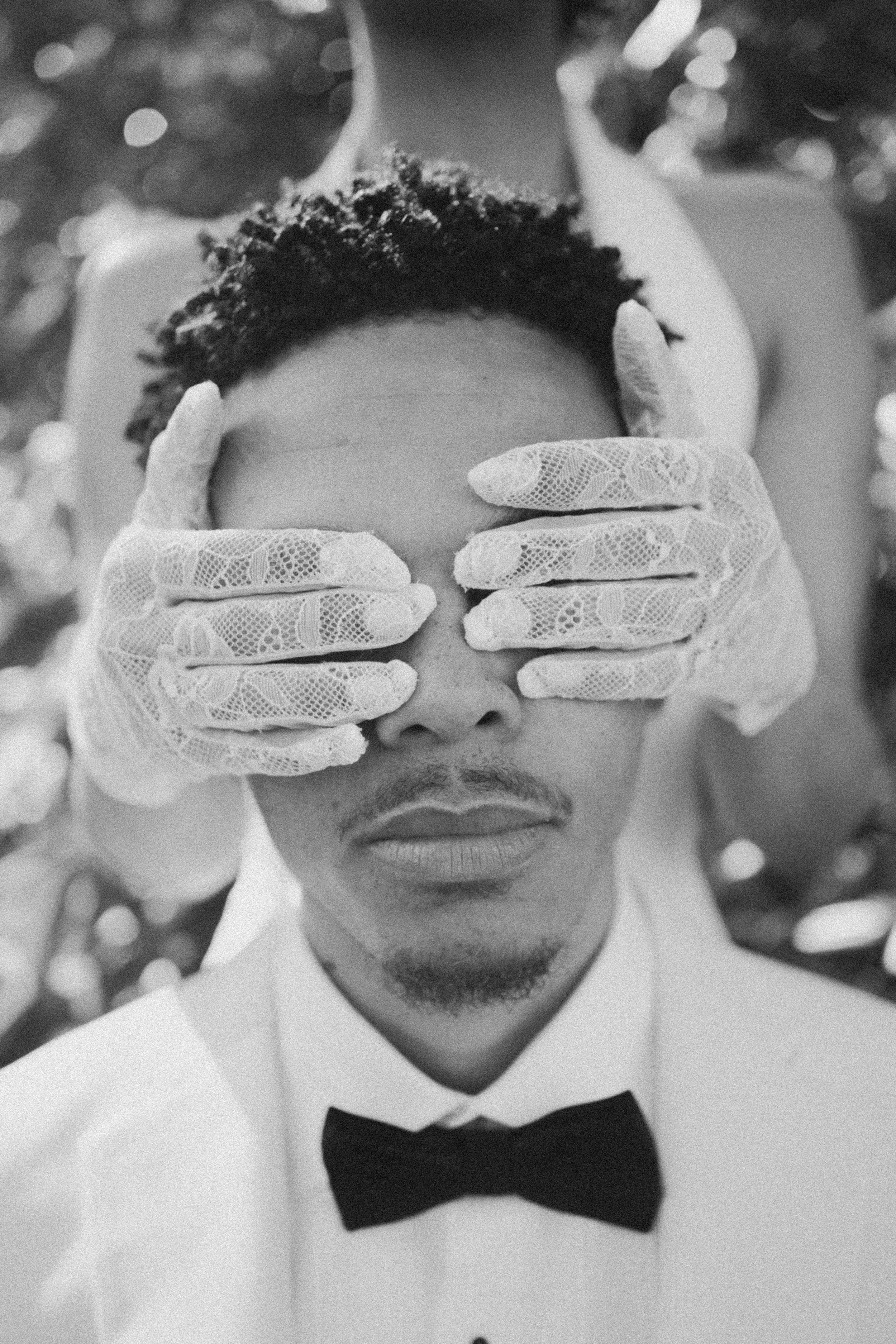 A man wearing a tuxedo, bowtie, and gloves, with his eyes covered by a person's gloved hands, appears to be in a formal setting with a glittery background.