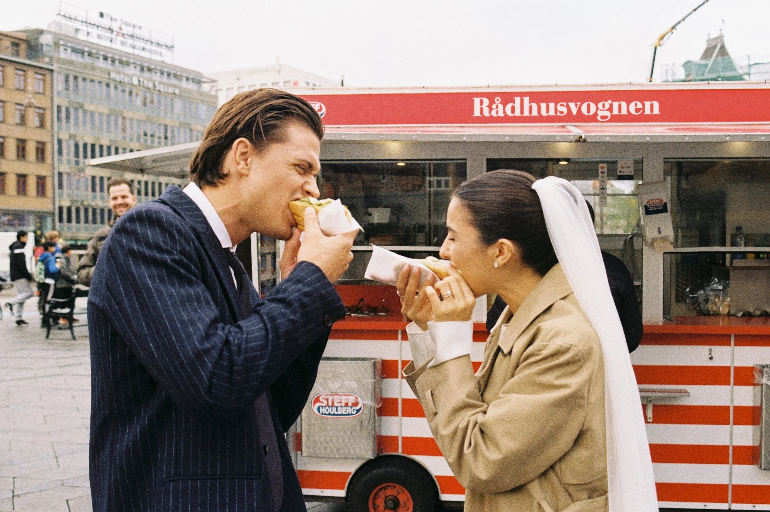 A man and woman sharing hot dogs at a street stand with a red sign that reads 'Rådhusvognen' in an urban public space.