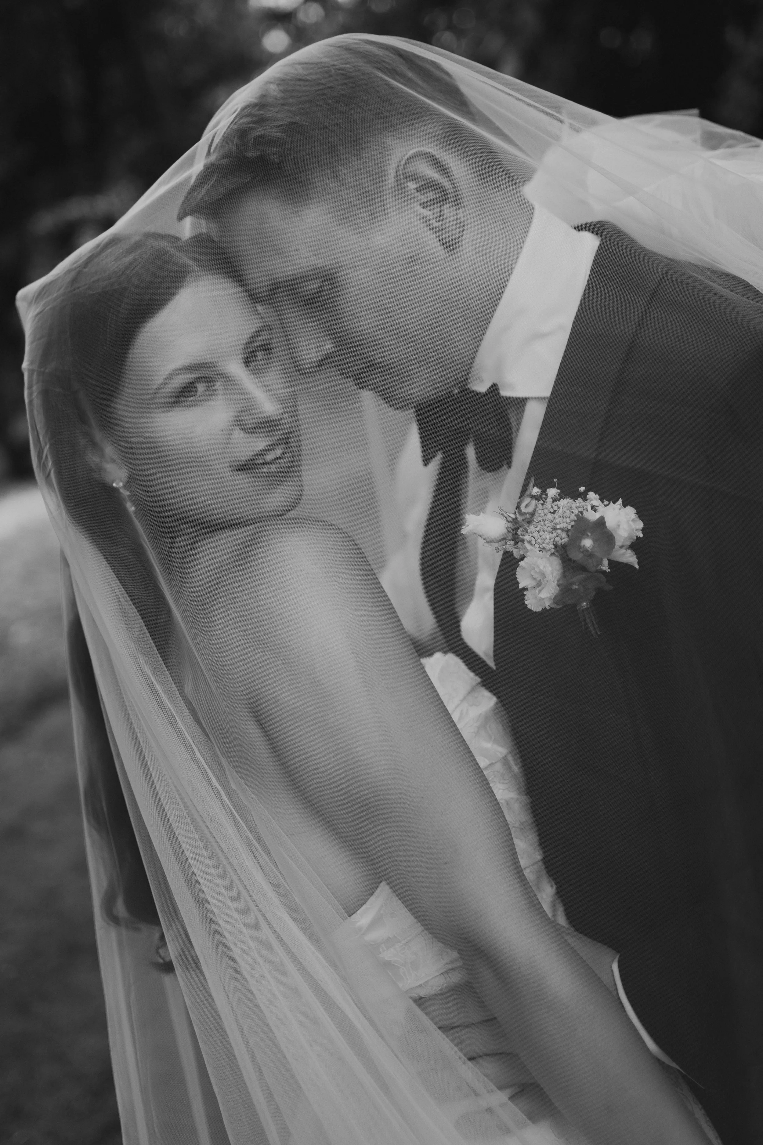 Black and white photograph of a bride and groom under a veil, close-up of their faces touching, outdoors background.