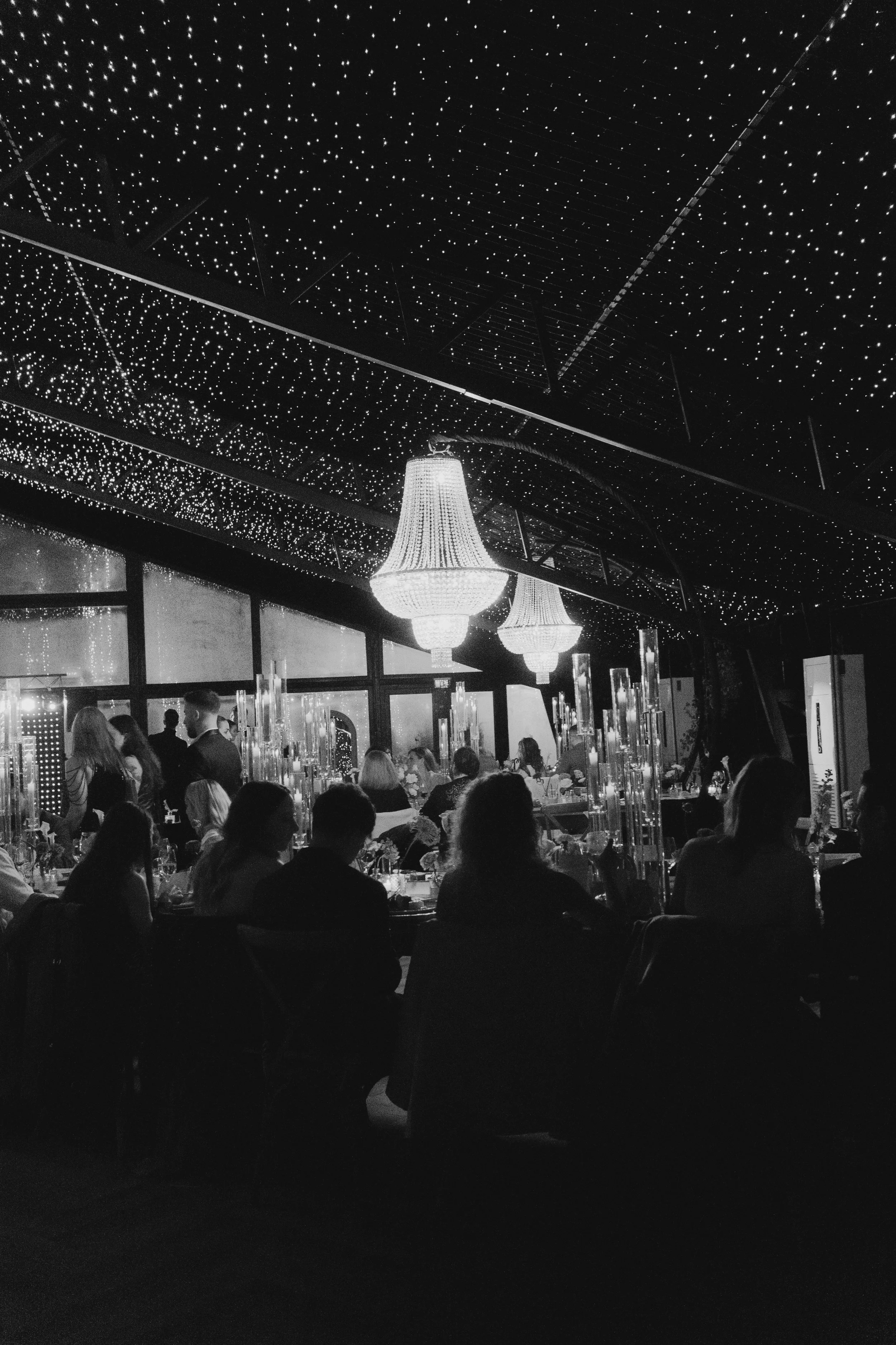 A restaurant or banquet hall filled with people sitting at tables, illuminated by elegant chandeliers, with a ceiling decorated with tiny lights creating a starry effect.