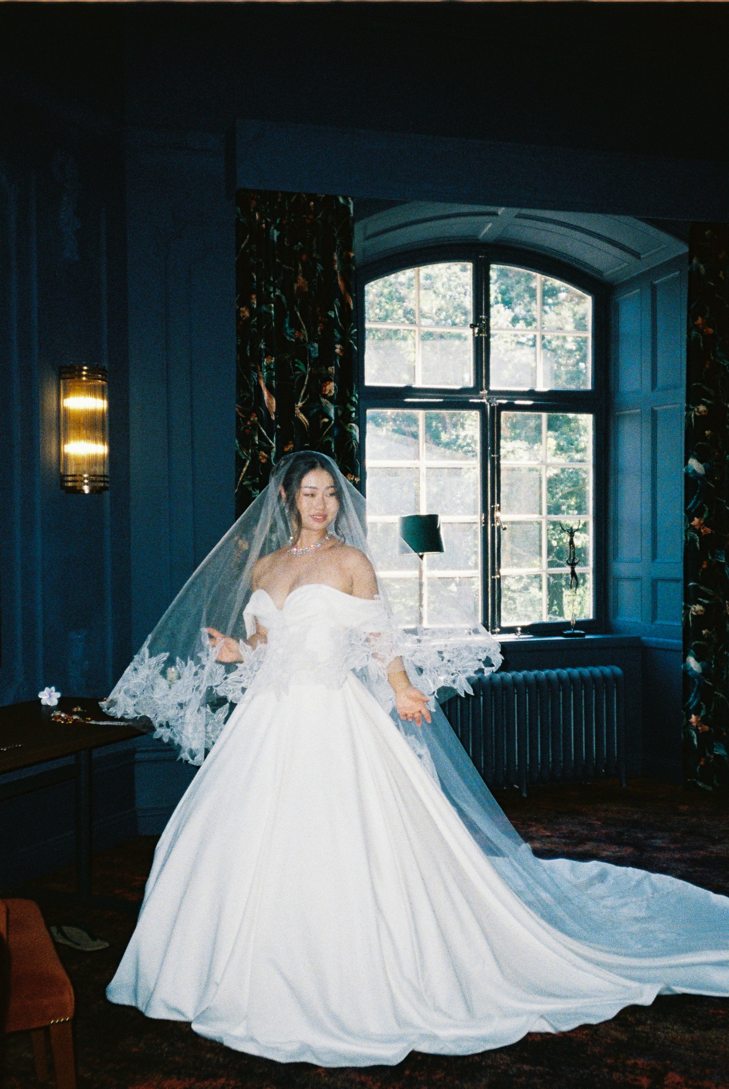 A woman in a wedding gown with lace details and a veil, standing indoors near a large window with floral curtains, in a dimly lit room with dark walls and a view of trees outside.