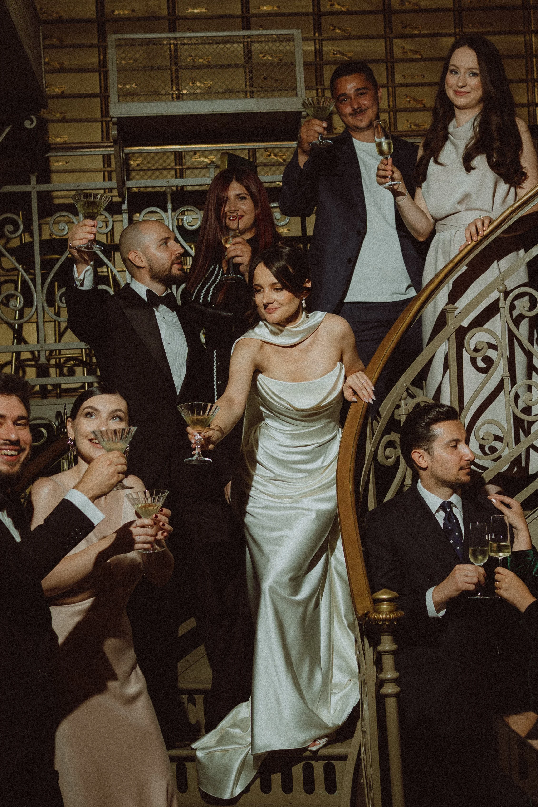 People dressed in formal attire celebrating with drinks on a staircase with ornate railing, in a luxurious setting.