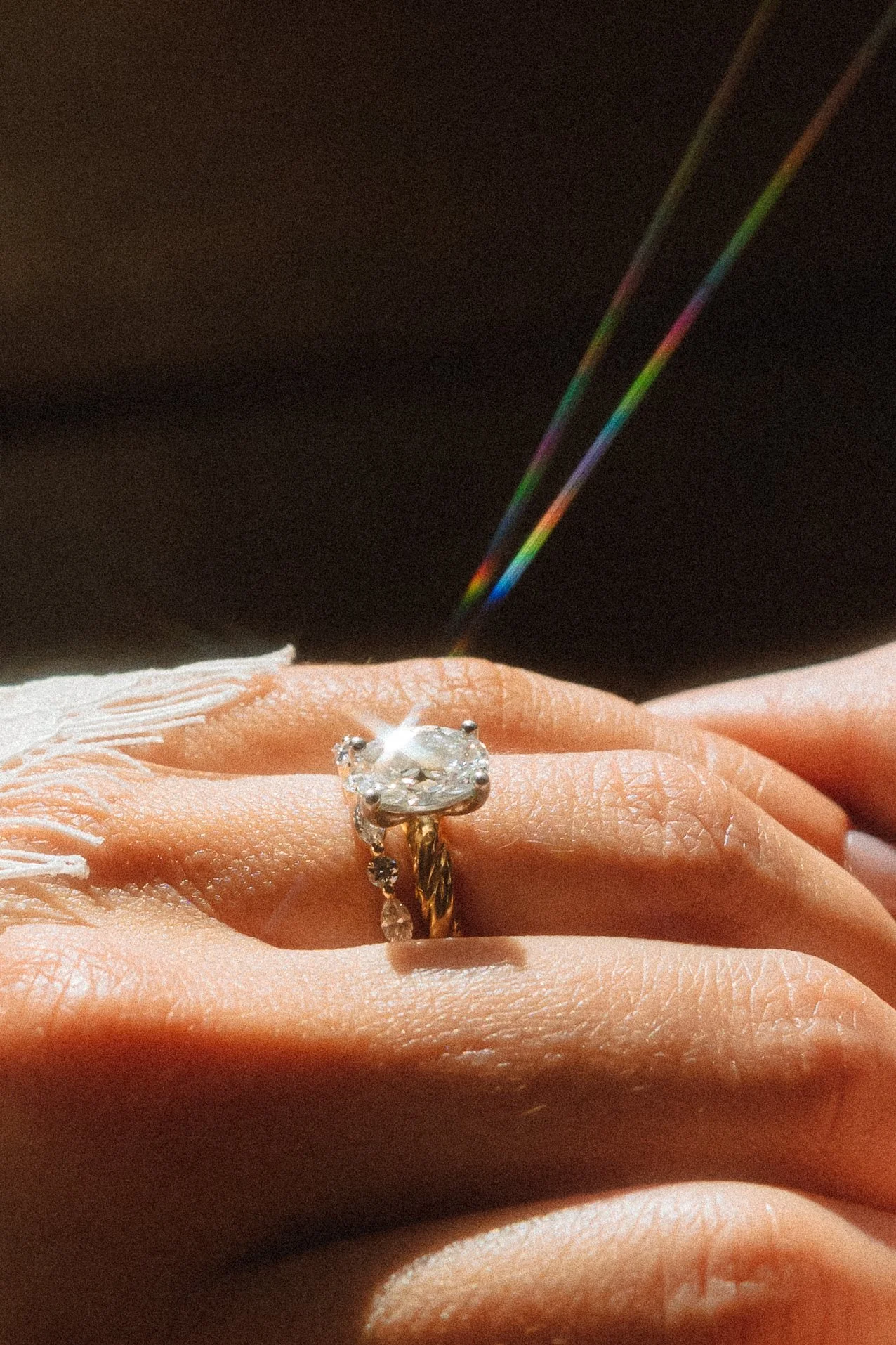 Close-up of a hand wearing a diamond engagement ring with a large center stone, on a person's finger, with rainbow light reflections in the background.