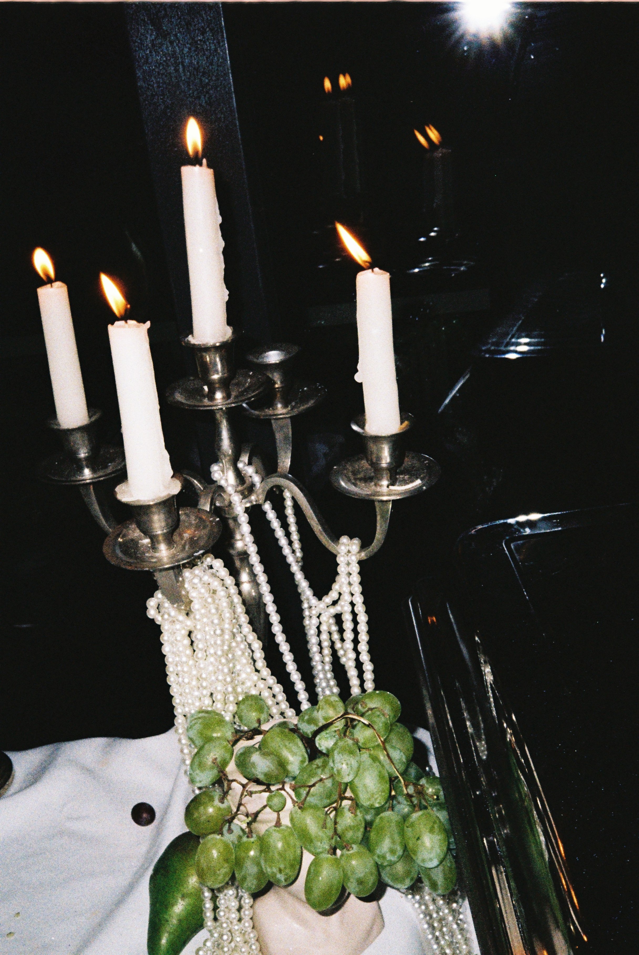 A metal candelabrum with four lit white candles surrounded by white pearl necklaces draped around it and a green vine plant at the base, set against a dark background with reflections.