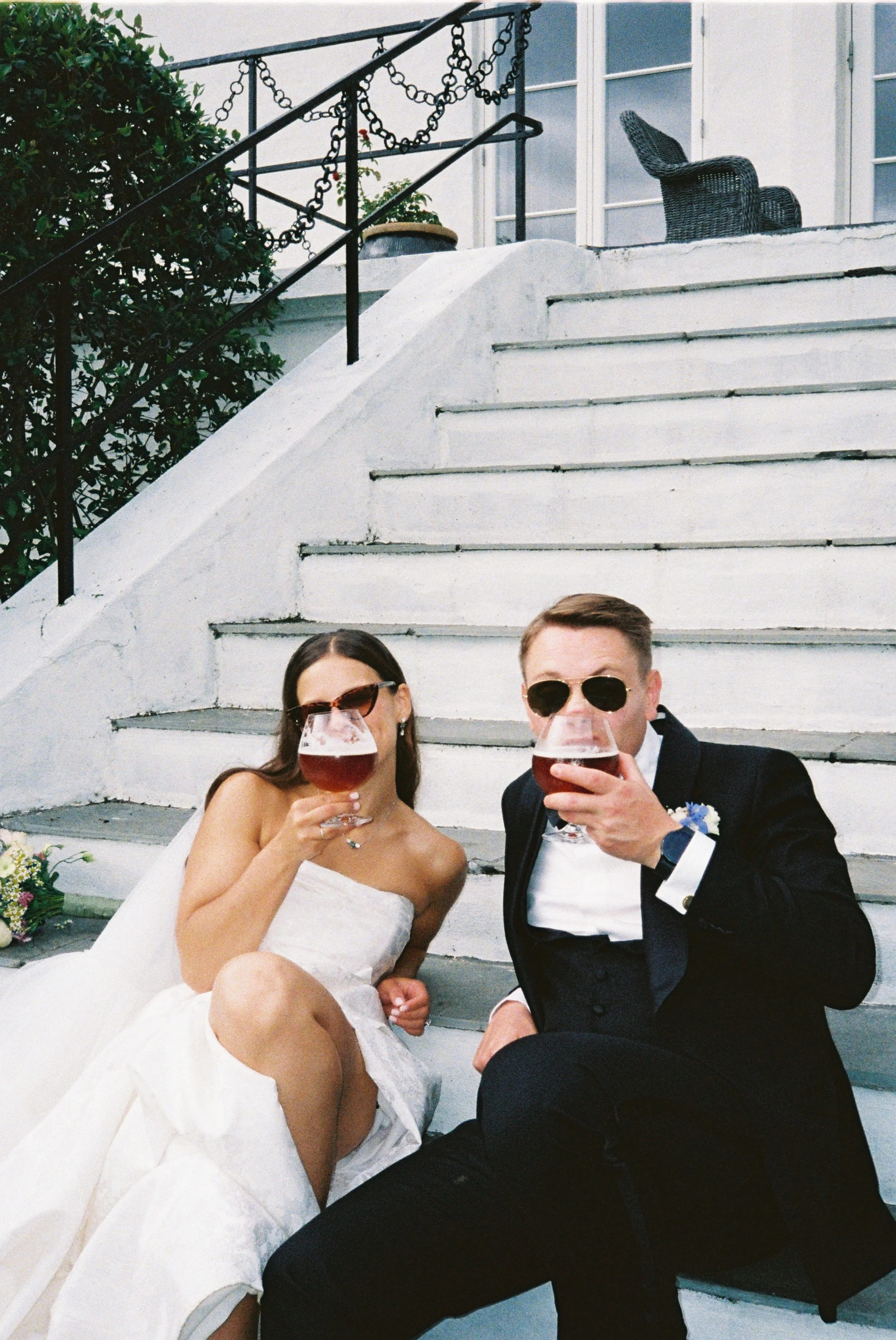 A bride and groom sitting on white stairs, wearing formal wedding attire, holding glasses of wine, and raising their glasses for a toast.