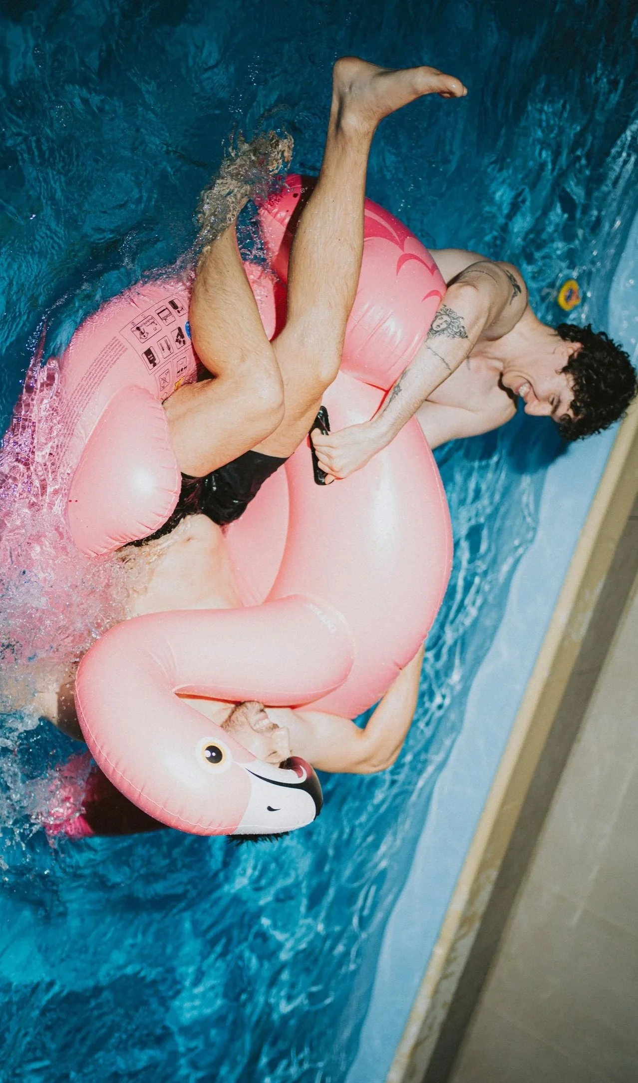 A man with dark curly hair and tattoos on his arm is lying on a pink flamingo pool float in a swimming pool, holding a remote control, smiling, with another person underneath him in the water.