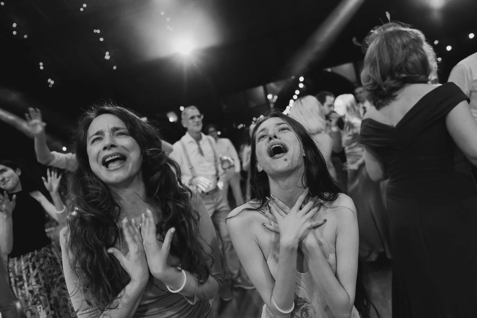 Two women singing with joy at a crowded event, with others around them singing or dancing.