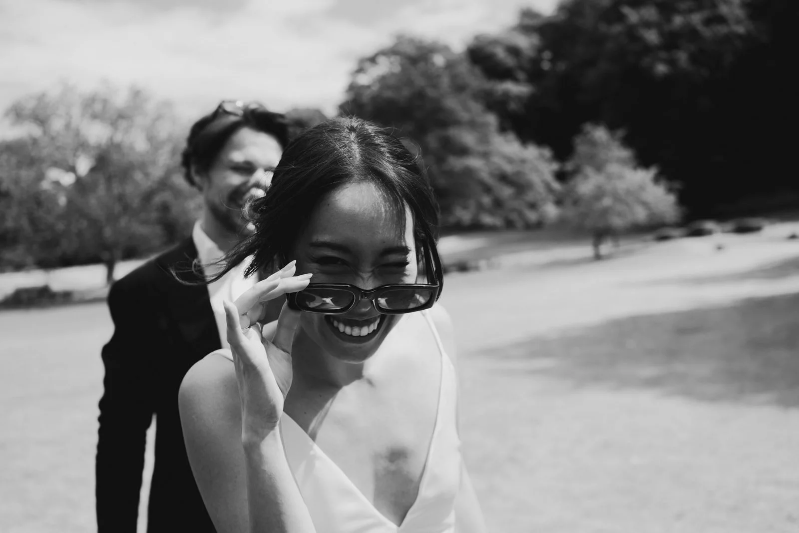 Black and white photo of two friends outdoors, smiling. The woman in the foreground lifts sunglasses and smiles, while the man in the background, slightly blurry, also smiles. Trees can be seen in the background.