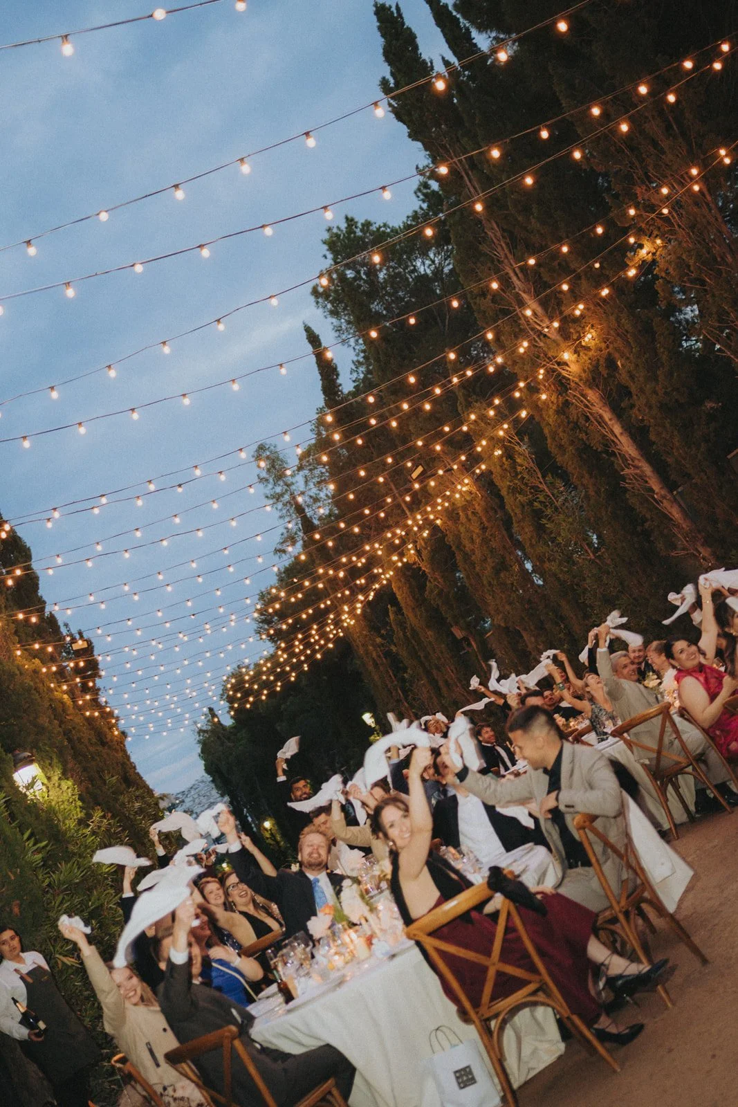An outdoor evening celebration under string lights, with people seated at long tables, waving white napkins, and enjoying food and drinks.