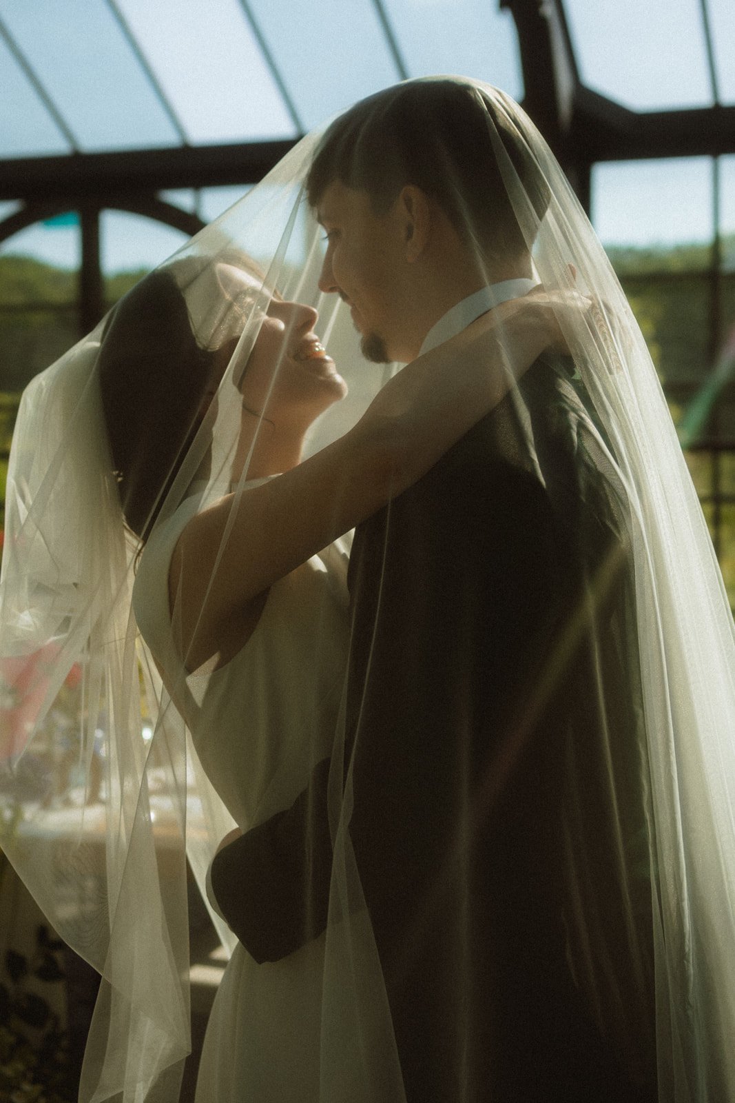 A bride and groom are dancing closely under a glass ceiling with sunlight streaming in, sharing a romantic moment.