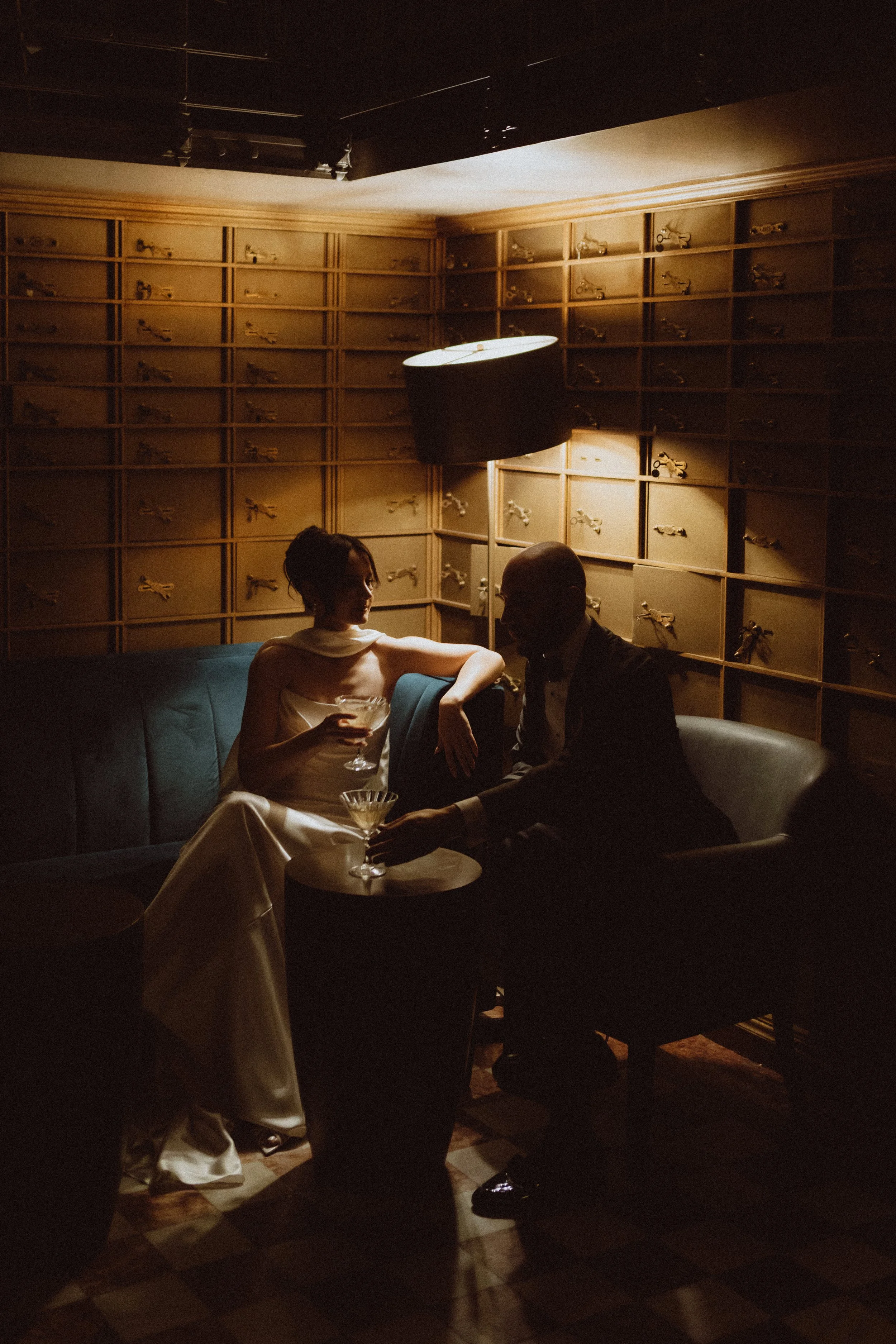 A couple dressed in formal attire sitting on a sofa in a dimly lit room with a wooden-paneled wall with multiple small drawers and vintage keys displayed on it. The woman is holding a cocktail, and they are engaging in conversation.