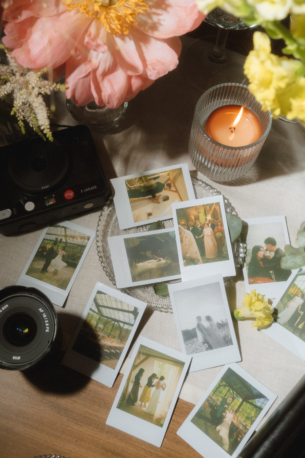 A table decorated with pink and yellow flowers, a lit candle in a glass holder, several vintage-style photographs, a camera, and a small vase.