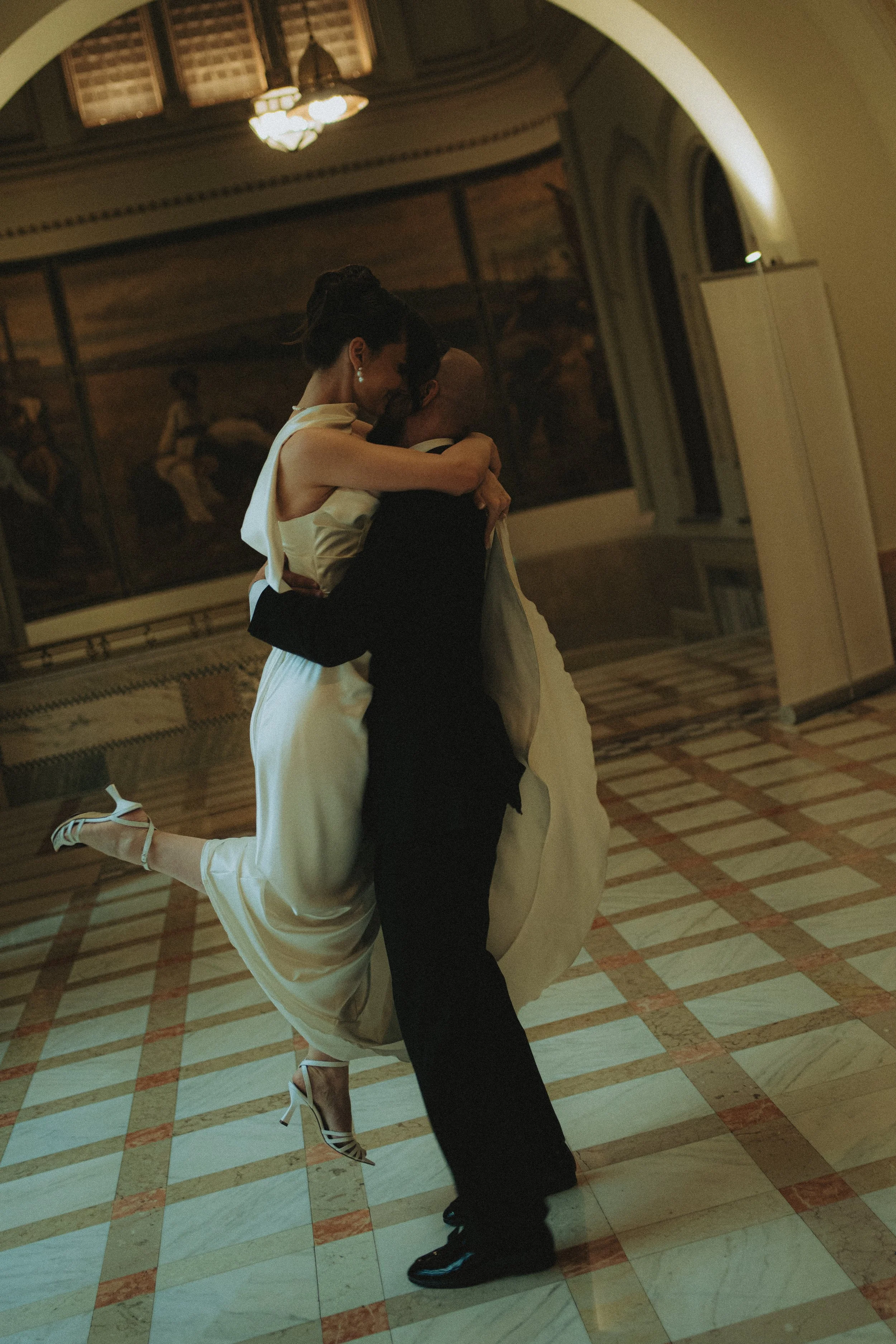 A man and a woman dance closely in an elegant, warmly lit room with ornate arches and a mural on the wall.