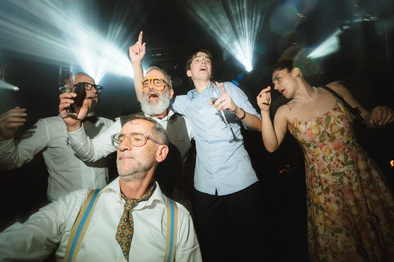 Group of people enjoying a celebration or party, dancing and holding drinks in a dimly lit room with colorful lights.