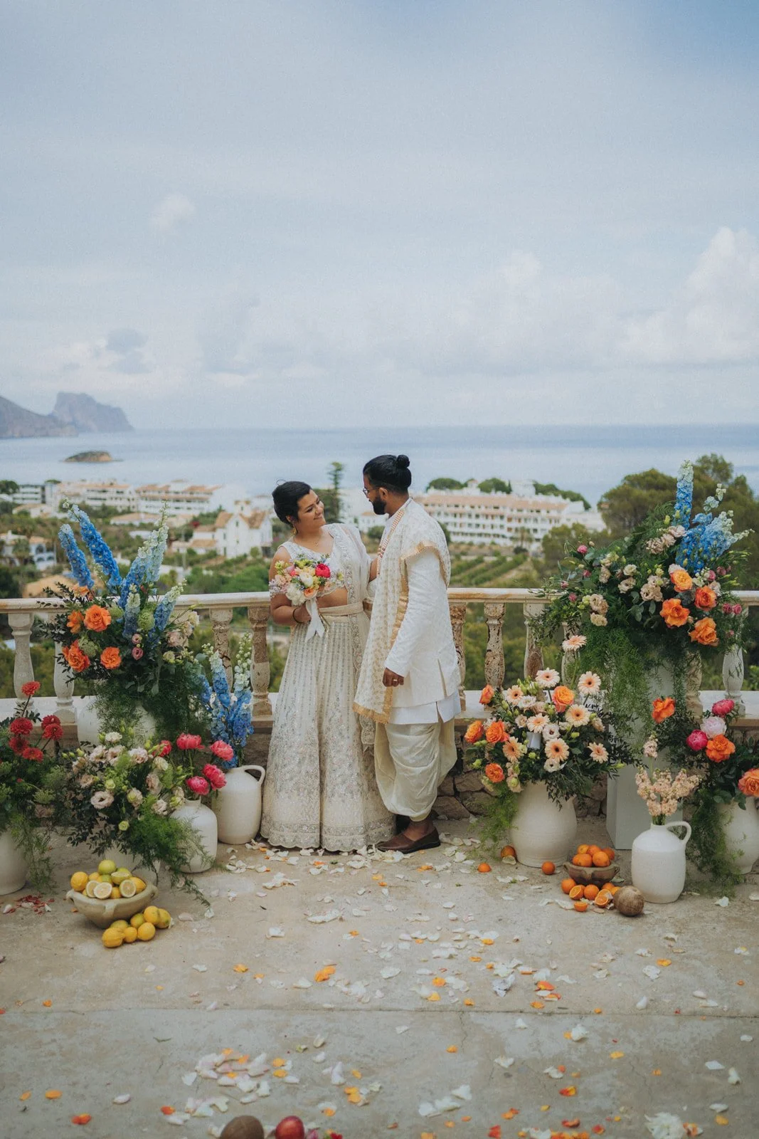 A couple dressed in traditional Indian wedding attire standing on a balcony decorated with colorful flowers. The woman holds a bouquet and the man looks at her. The background shows a scenic ocean view with some buildings and greenery.