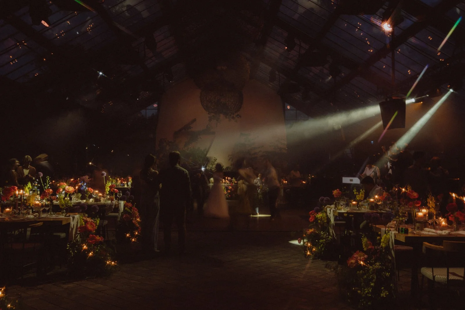 Dimly lit indoor event with people socializing, floral decorations, and dramatic stage lighting.