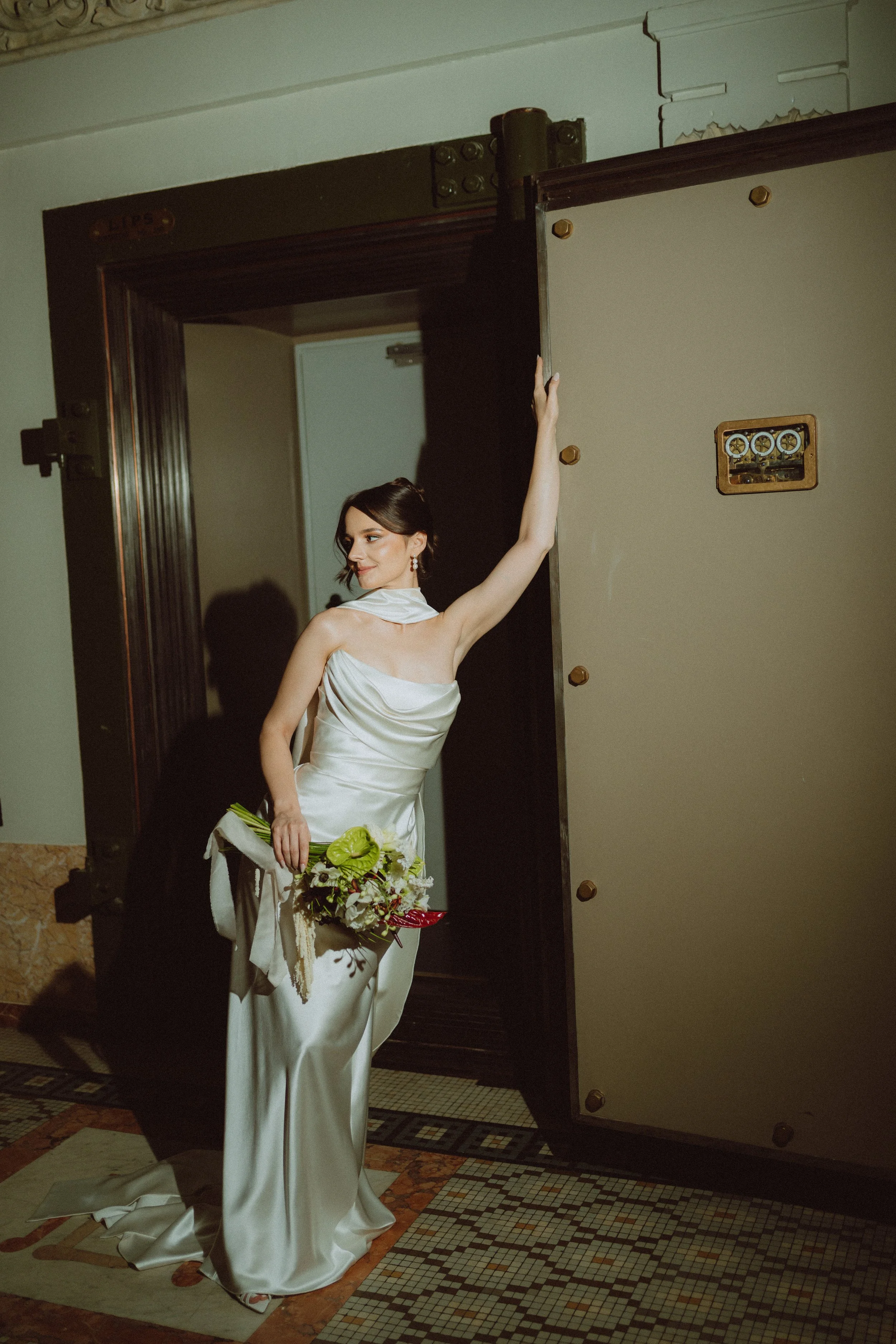A woman in a long white satin wedding dress holding a bouquet of white and green flowers, standing in a doorway with a dark wooden door and a beige wall.