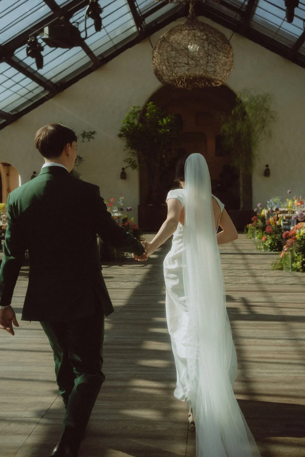 A bride and groom holding hands during their wedding ceremony in a decorated indoor venue with flowers, plants, and large windows.