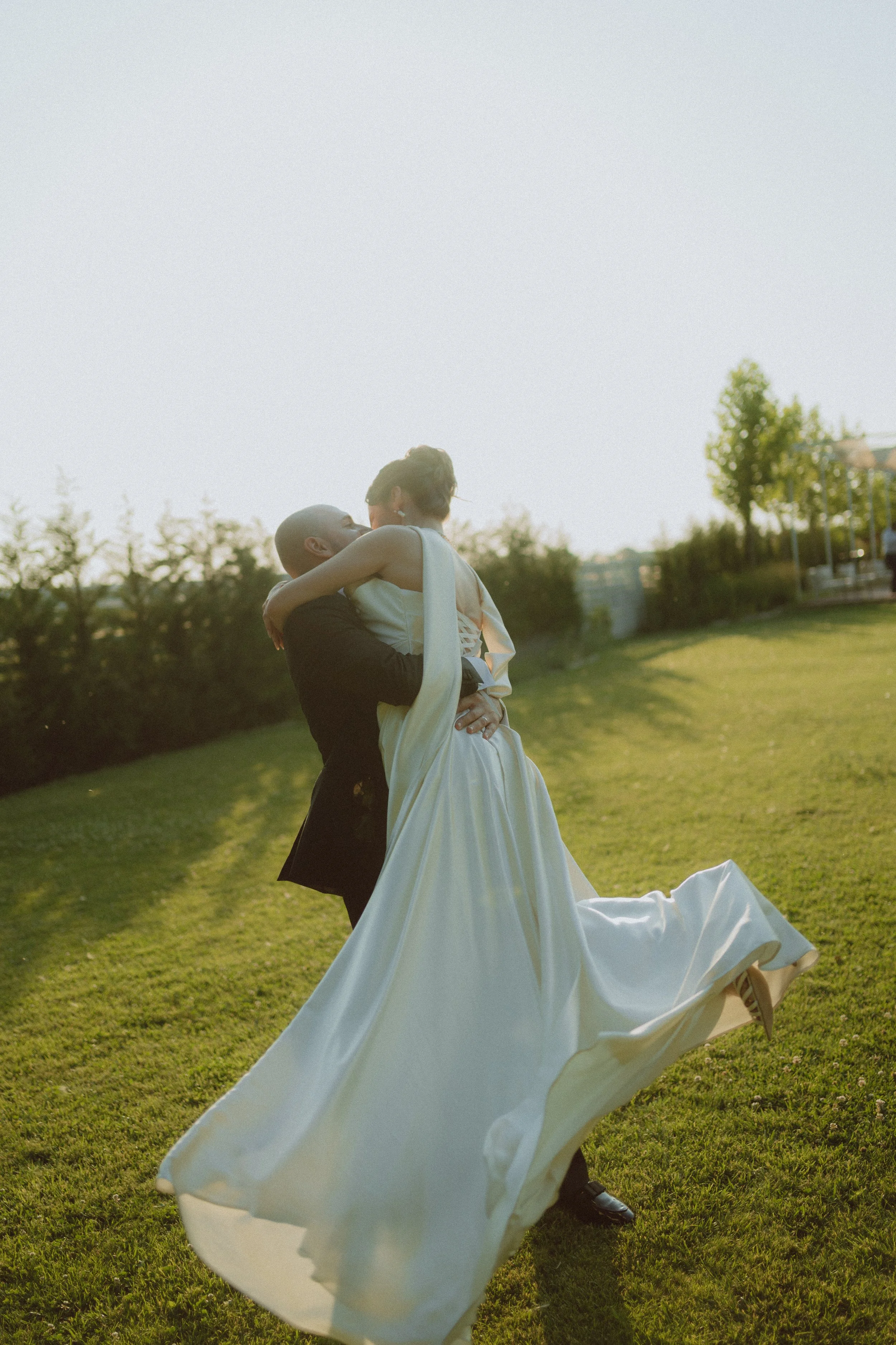 A bride and groom sharing a dance outdoors on a grassy field in the evening, with trees and a house in the background.