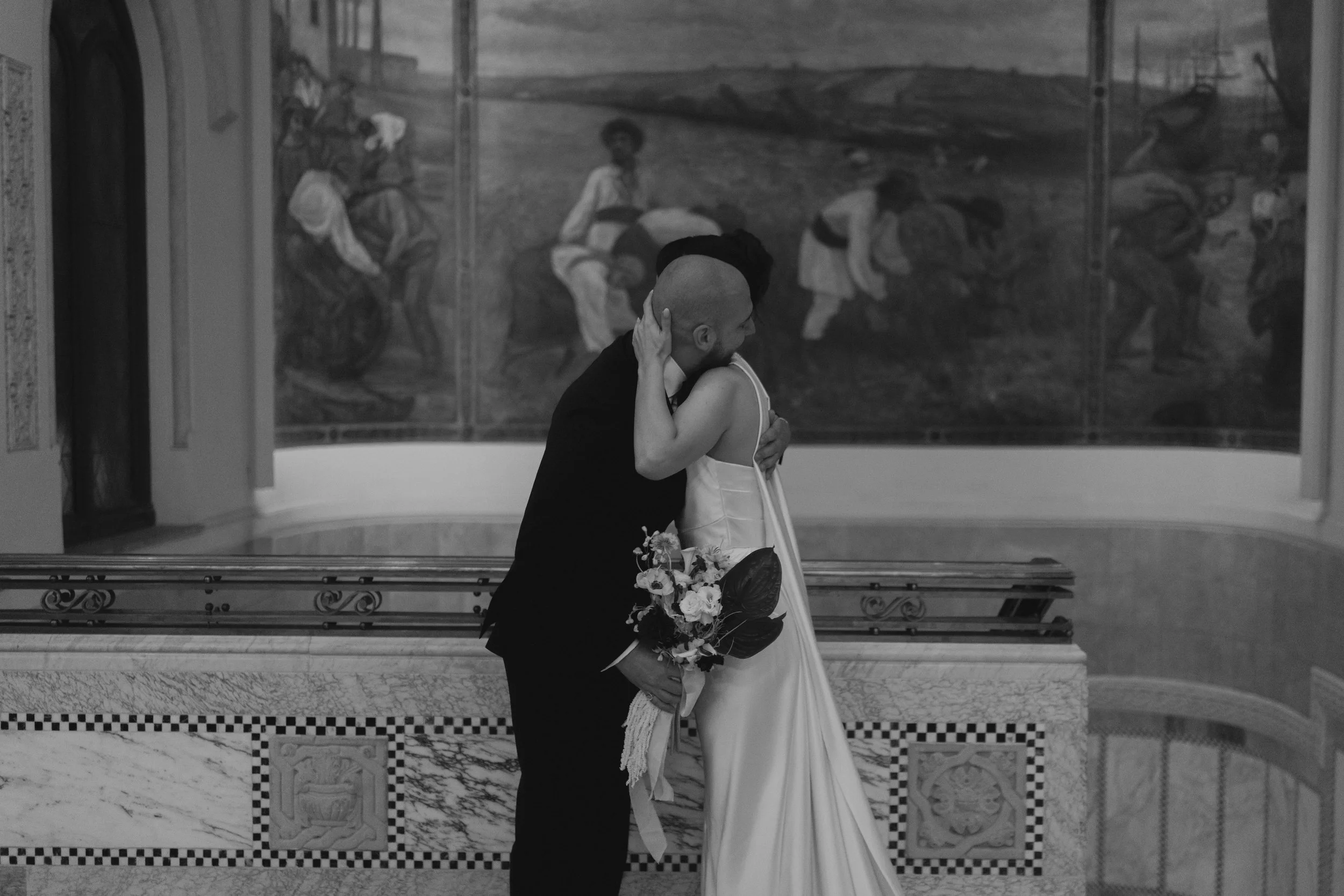 A couple dressed in wedding attire embracing each other indoors with a large painting in the background and marble flooring.