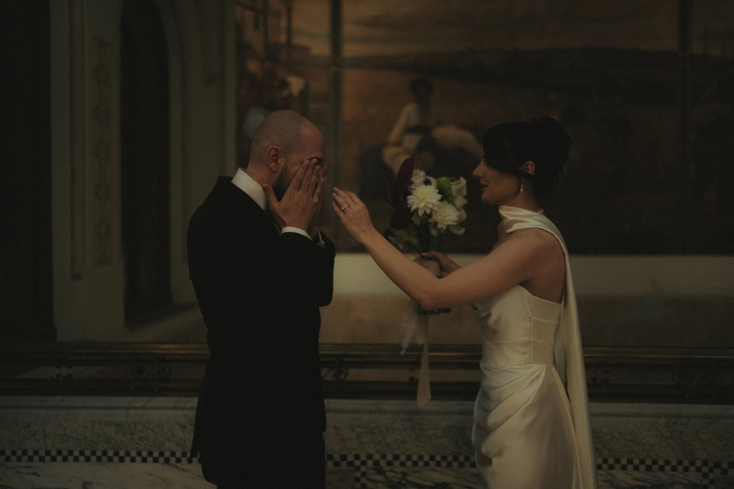 A woman in a satin dress presents a bouquet of flowers to a man in a tuxedo, with another woman in the background observing, set in an elegant indoor space.