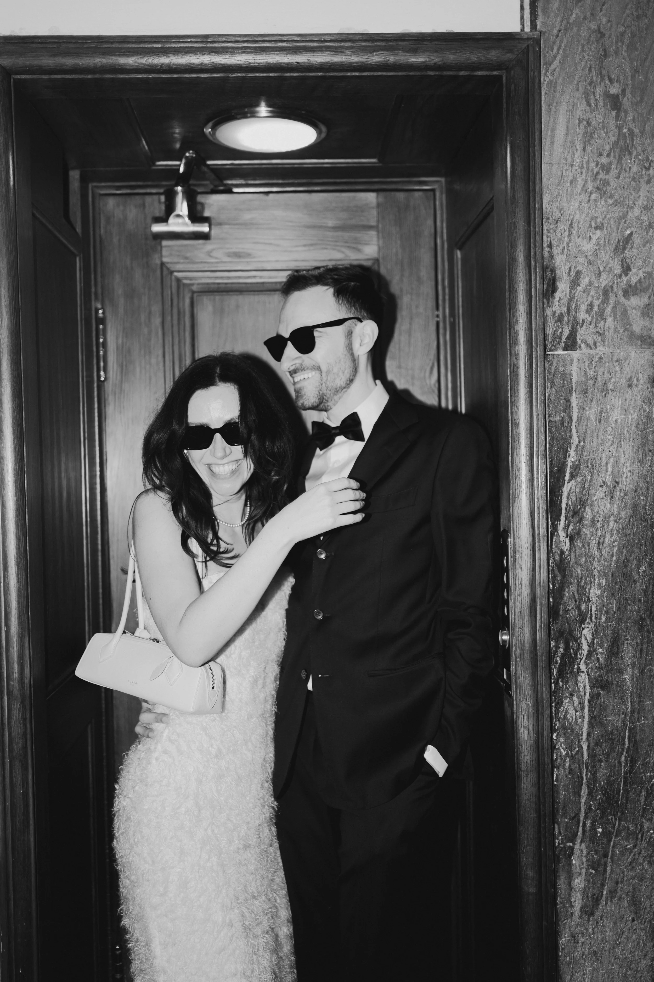 A black-and-white photograph of a smiling woman and man in formal attire, wearing sunglasses, posing together in an elevator with wooden walls and a marble wall to the right.