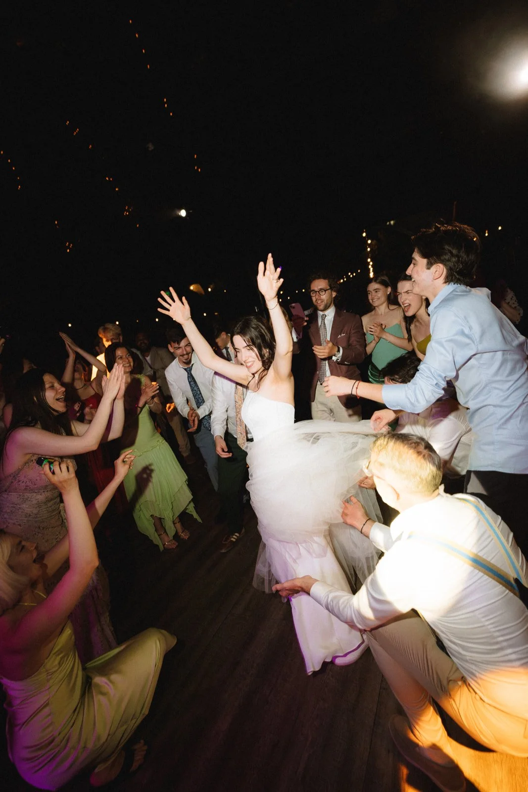 People dancing and celebrating at a wedding reception under string lights at night.
