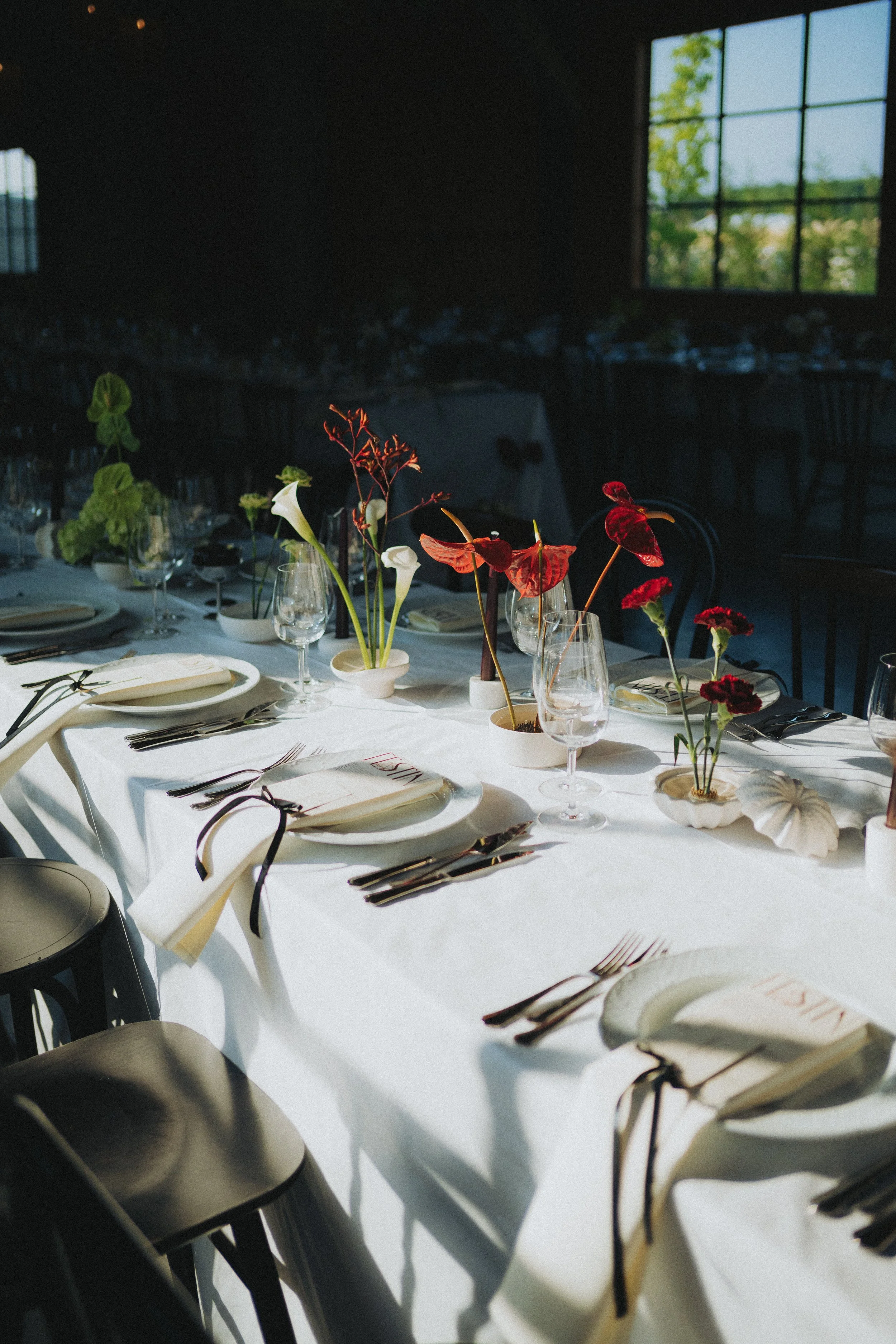 A long banquet table with a white tablecloth, set with plates, silverware, wine glasses, and place cards. Small vases with colorful flowers decorate the table. The room has dark walls and large windows with a view of trees outside, allowing natural l