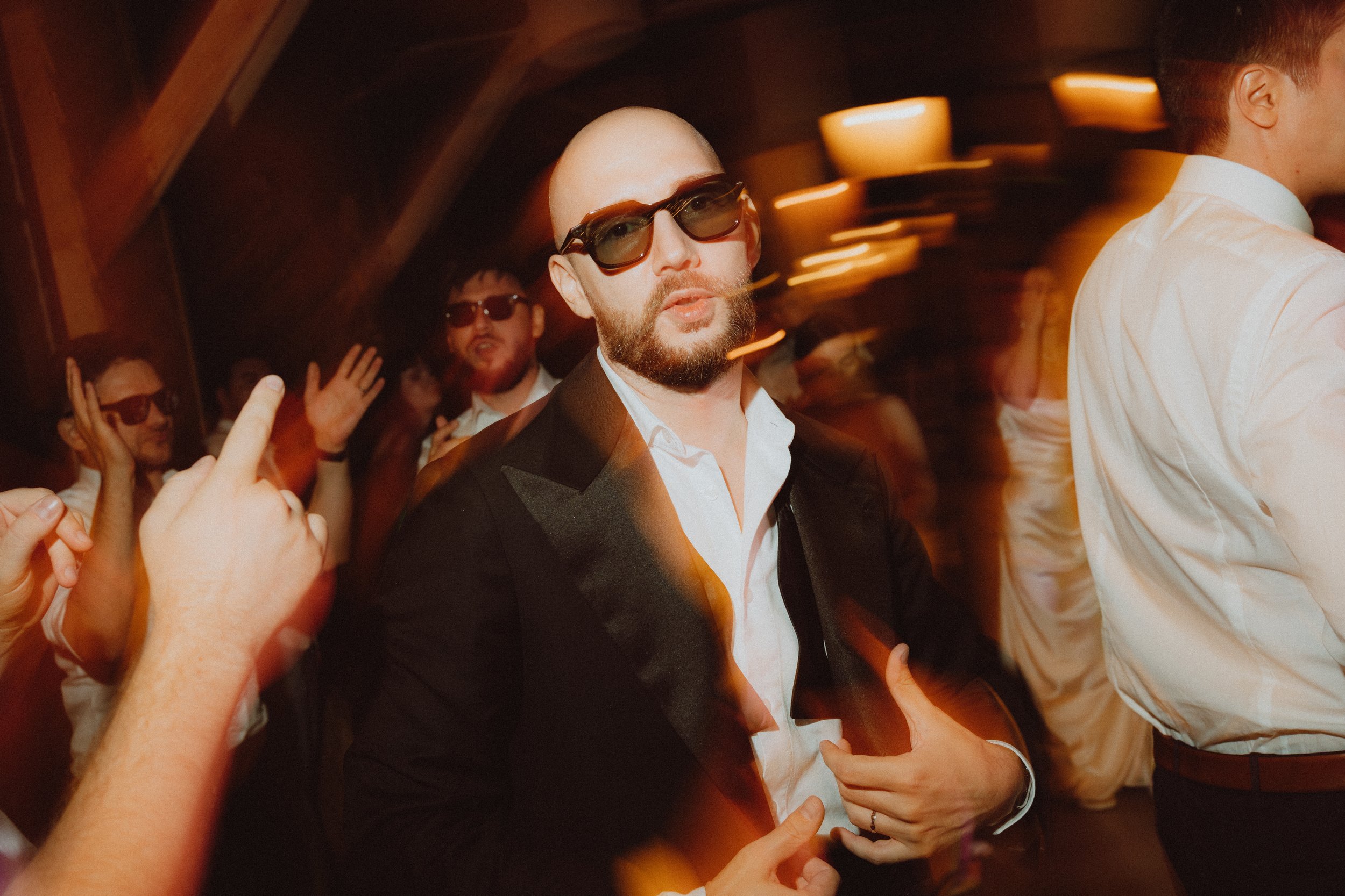 A group of people at a social gathering, with one man in the center wearing sunglasses and a black suit with a white shirt, surrounded by others in casual and semi-formal attire, some wearing sunglasses, in a warmly lit setting with motion blur effec