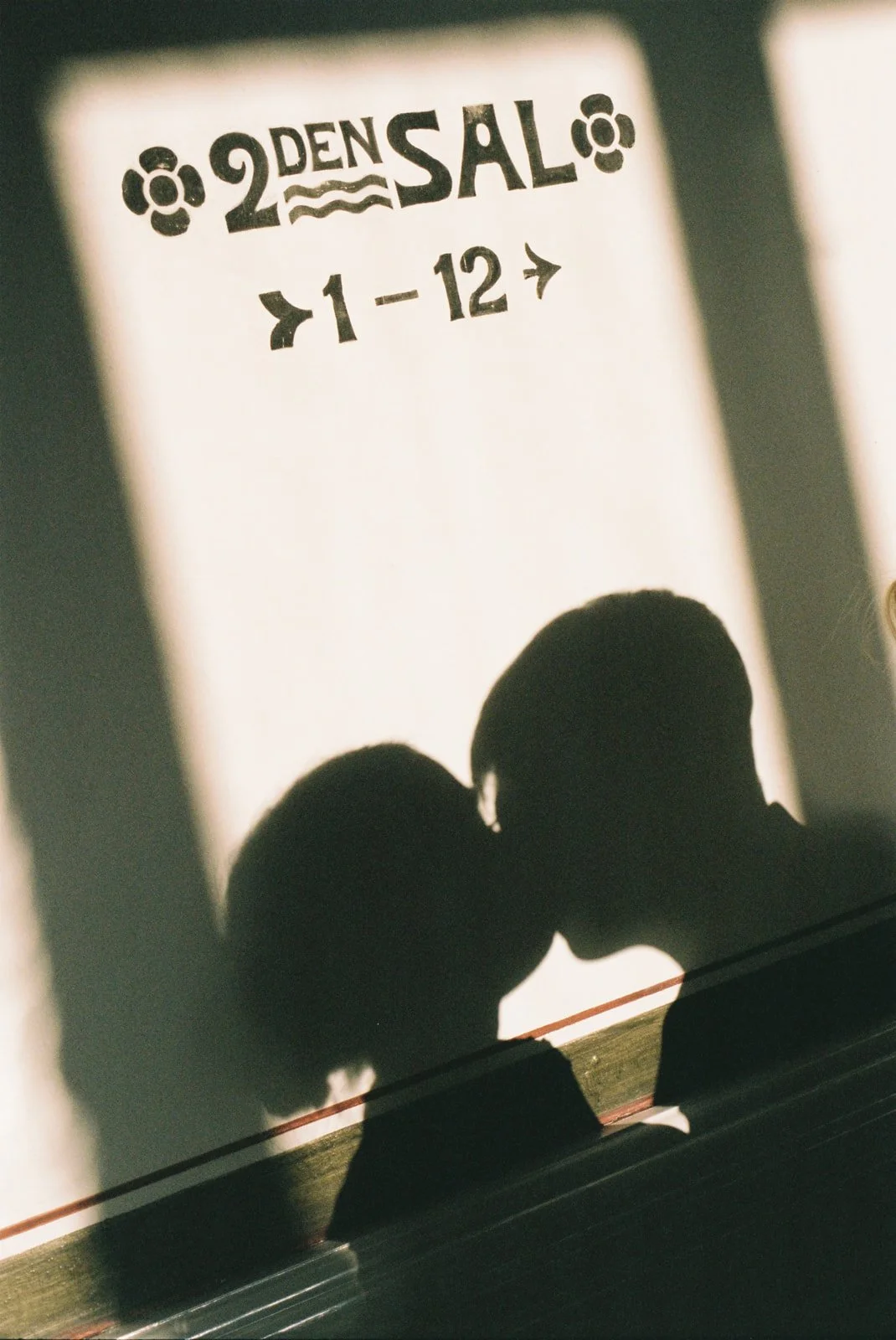 Shadow of two people kissing on a wall with a sign that reads ''DENS AL 2'' and arrows pointing left and right, and numbers ''1-12''.