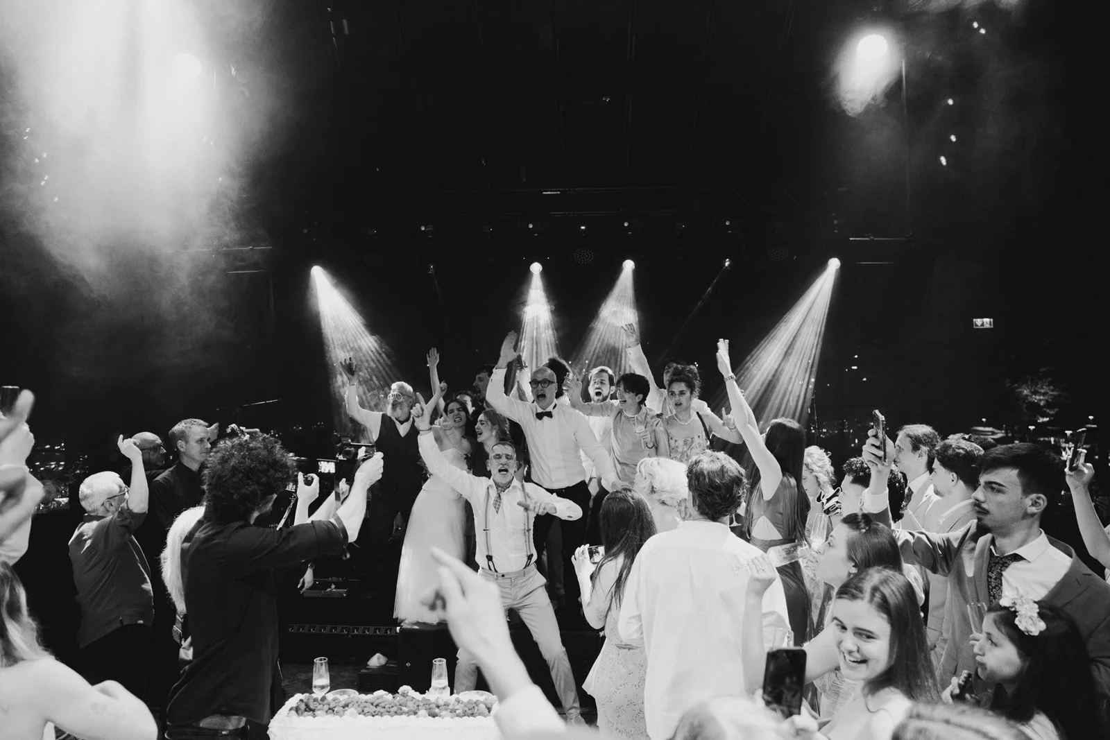People celebrating at a party with some on a stage, with spotlights shining down and a cake in the foreground.