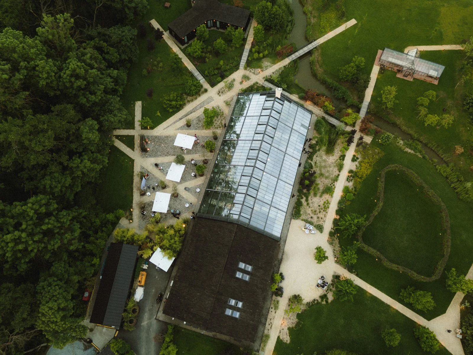 An aerial view of a garden area with a large glass conservatory building, pathways, outdoor seating with umbrellas, and lush green trees and landscape.