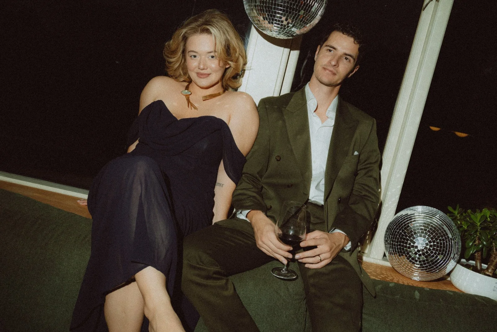 A woman with shoulder-length red hair sitting closely next to a man with dark curly hair near a window at night. The woman is smiling and wearing a dark off-the-shoulder dress, and the man is holding a glass of red wine and dressed in a green suit. There is a disco ball hanging above them and a speaker on the floor nearby.