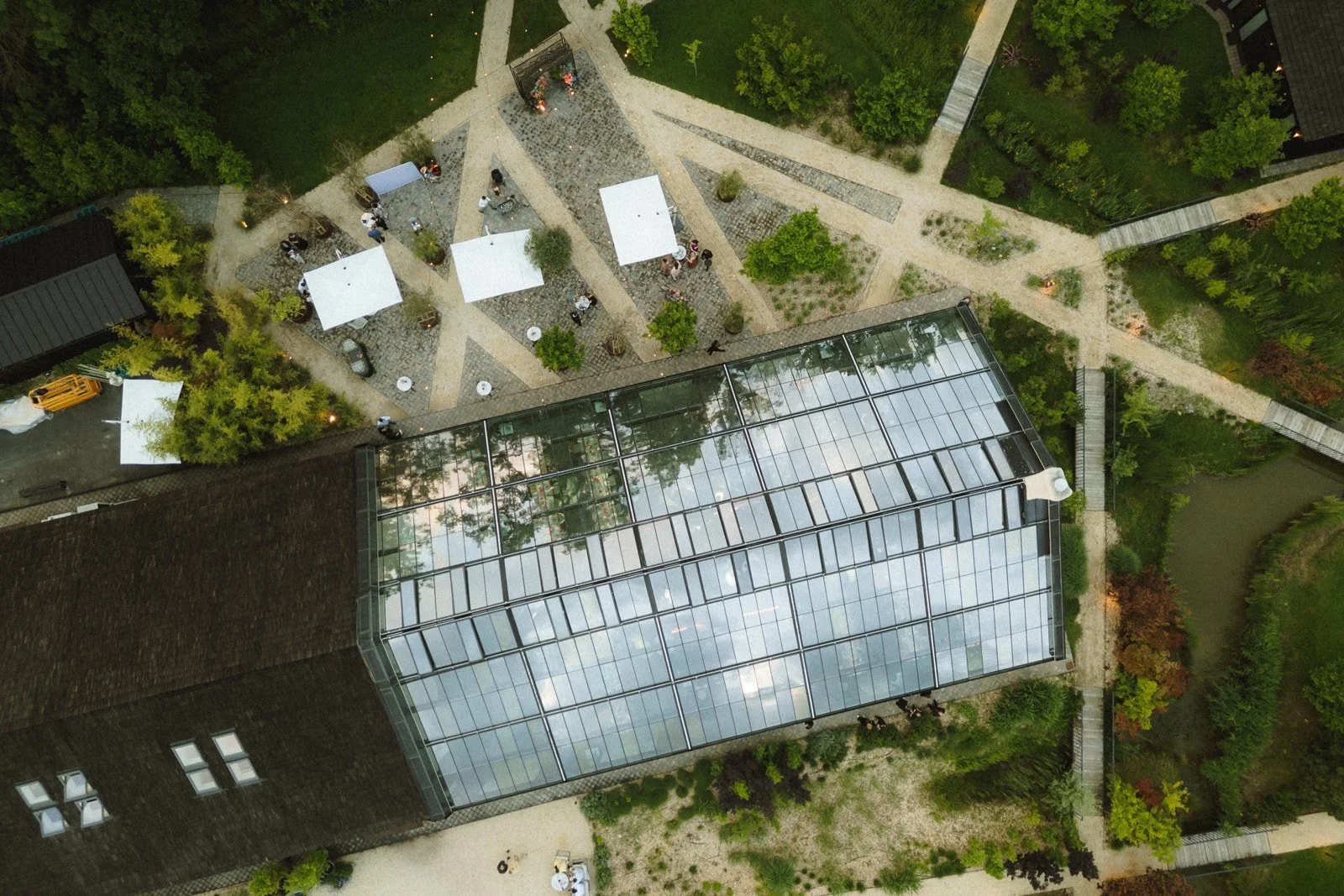 An aerial view of a garden with paved walkways, green trees, small tables with umbrellas, and a glass greenhouse attached to a building.