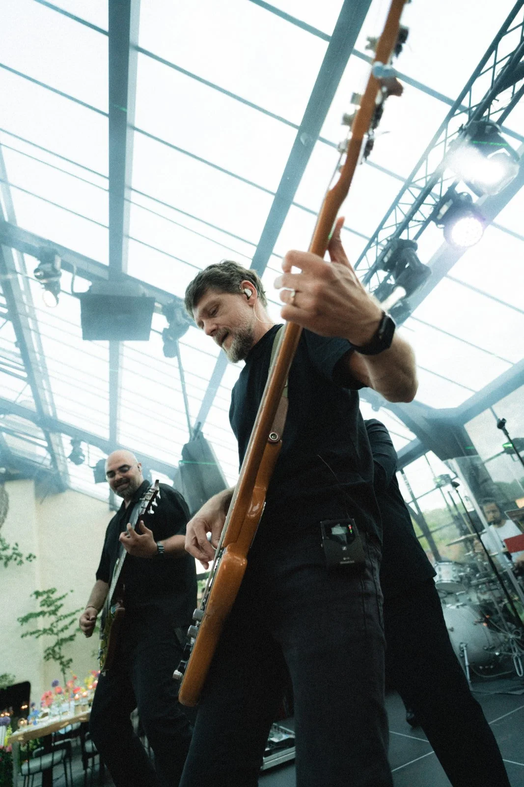 Two musicians playing guitars on stage under a glass roof with lighting equipment overhead.