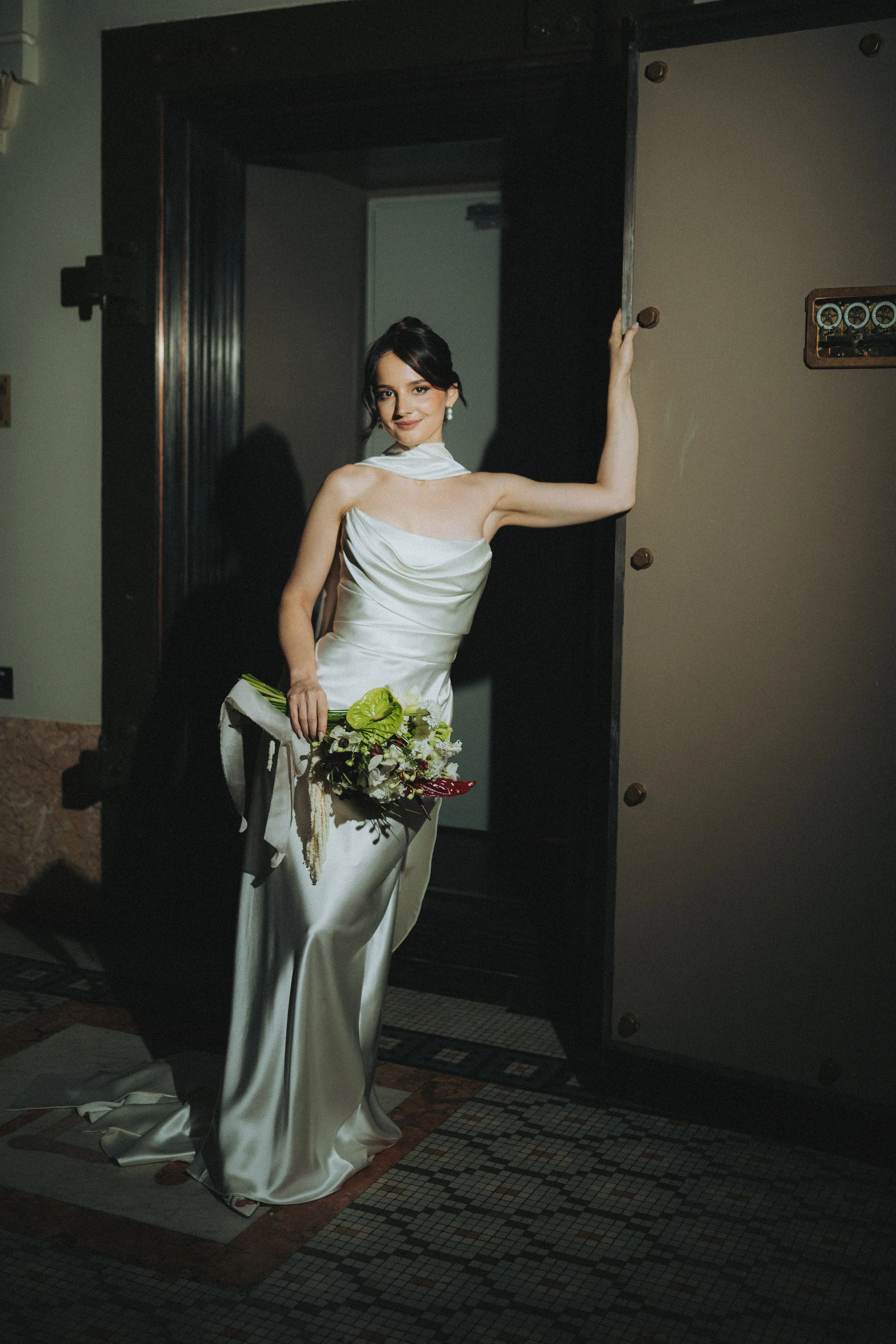 A woman in a white dress holding a bouquet of flowers, standing in front of a black door with a modern design, inside a building with a patterned tiled floor.