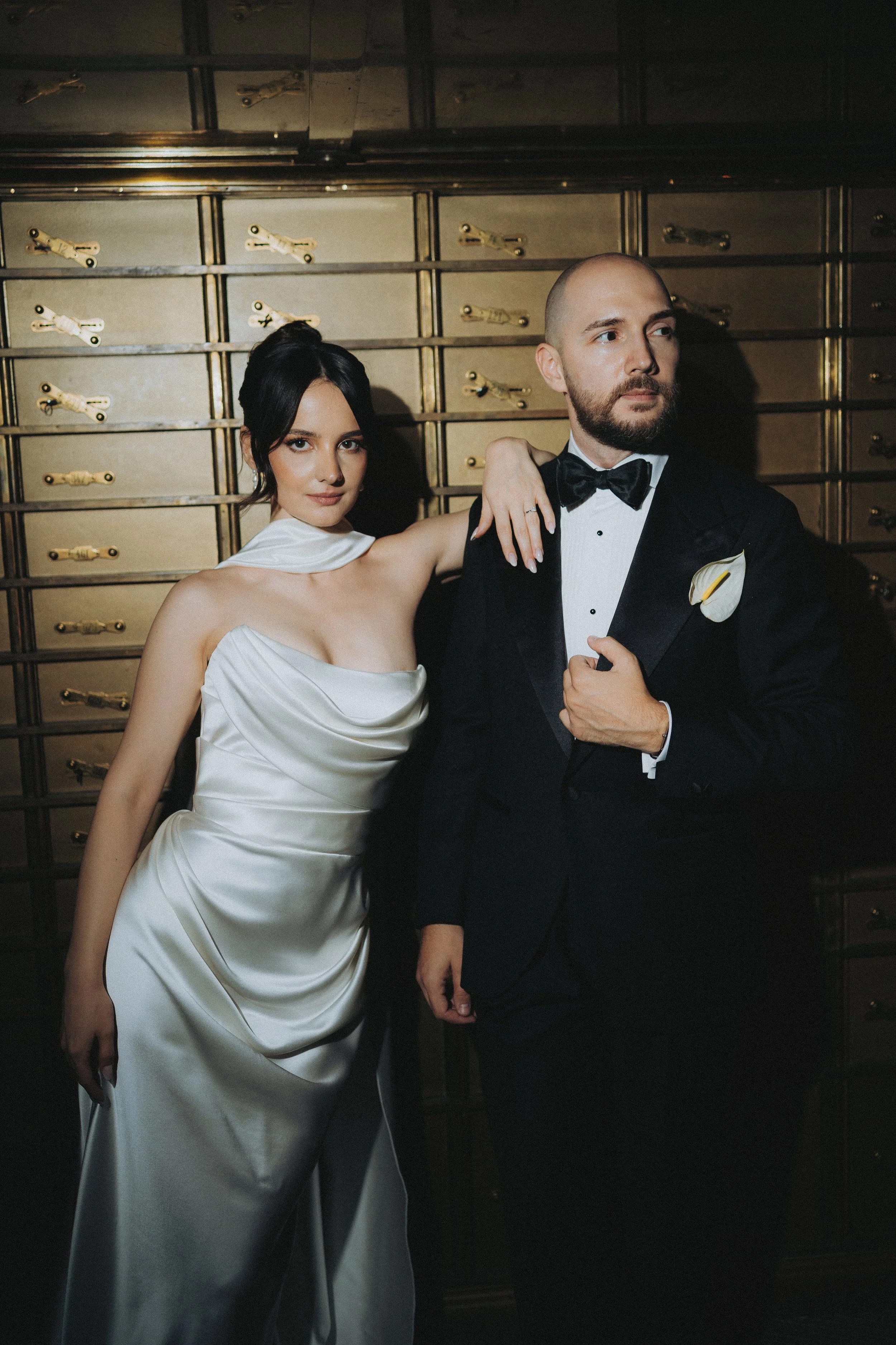 A woman in a white satin gown and a man in a black tuxedo with a bow tie are posing together in front of a wall of gold-colored drawers. The woman's arm is draped over the man's shoulder, and the man's arm is crossed over his chest. The woman has dar