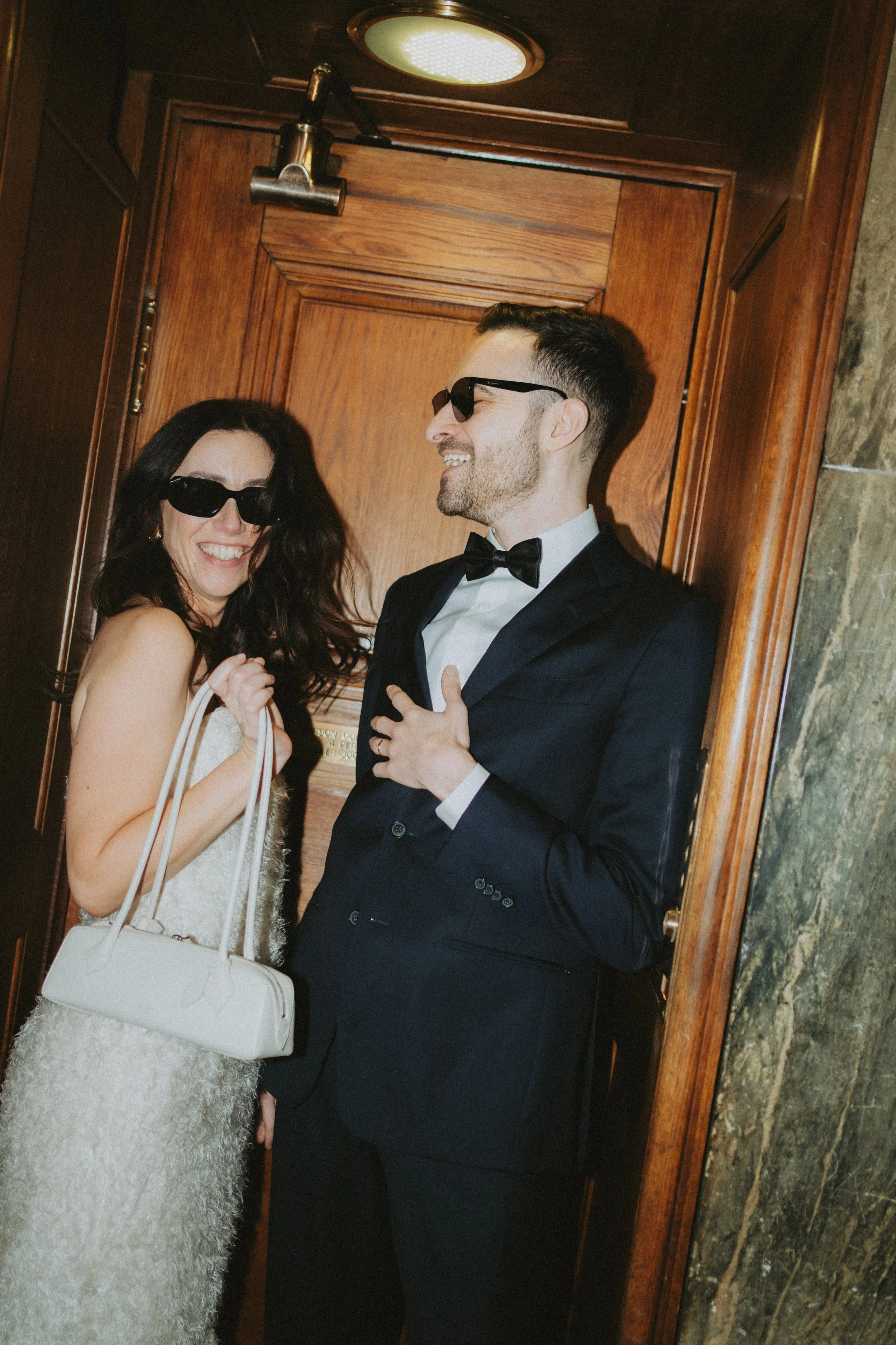 A woman and a man, both wearing sunglasses, are smiling and looking at each other in an elevator with wooden doors. The woman is holding a white purse and has long dark hair, while the man is dressed in a tuxedo with a bow tie and has short dark hair