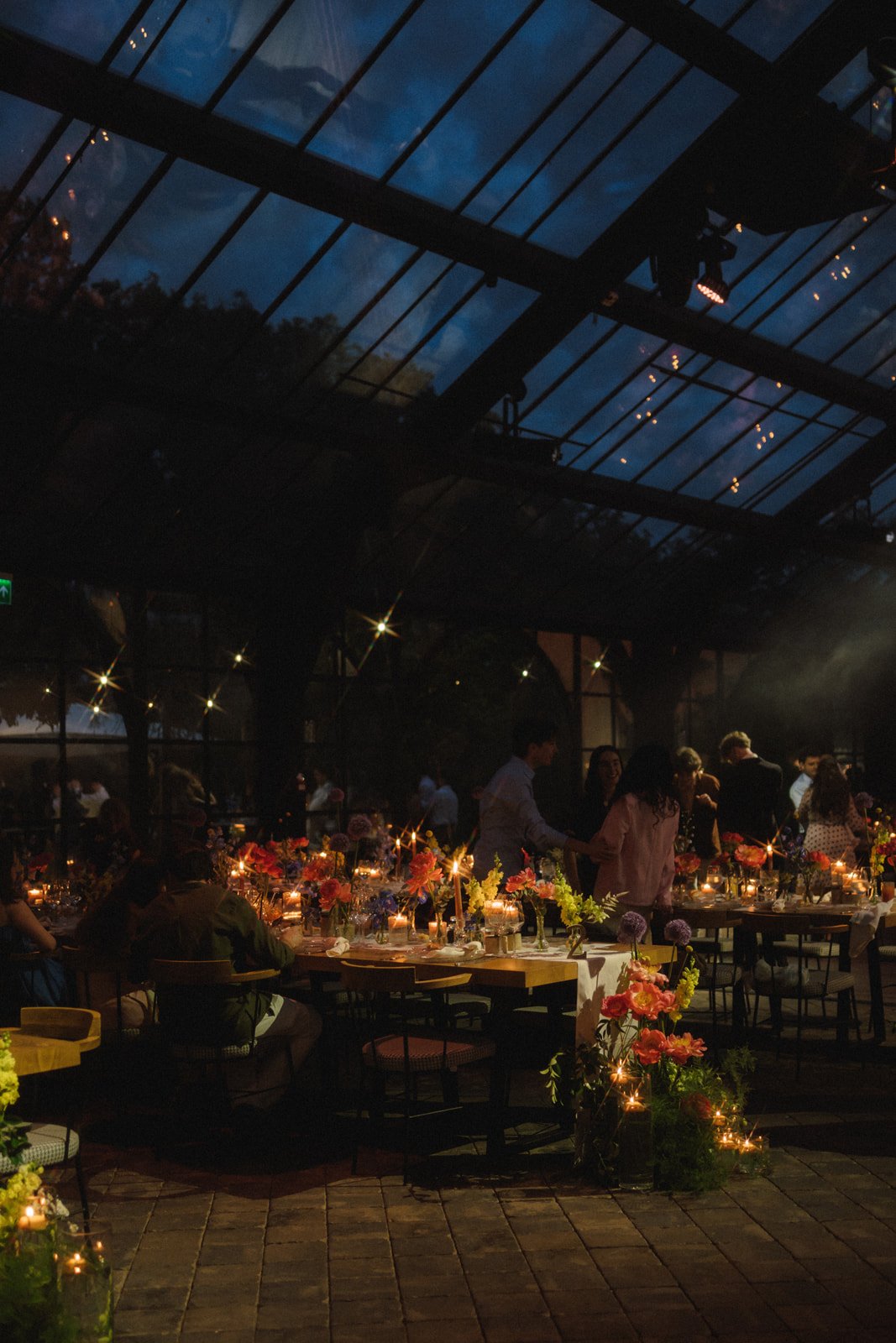 A dimly lit event space with tables decorated with flowers and candles, filled with people socializing, under a large glass ceiling showing the evening sky.
