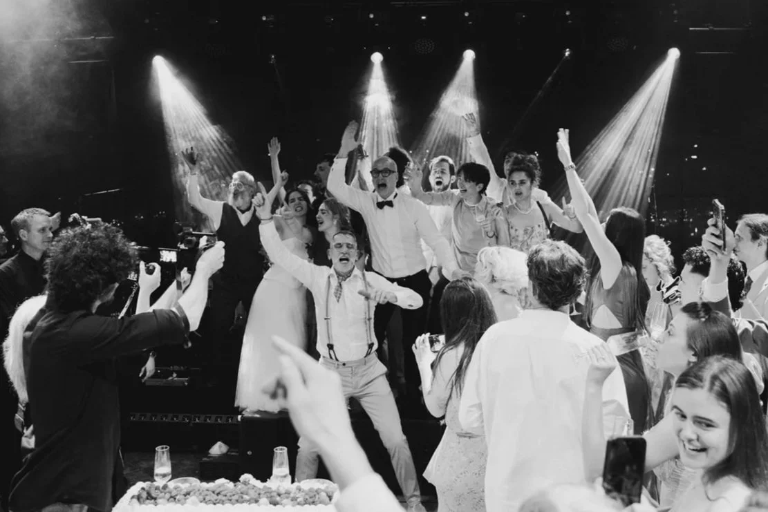 Group of people celebrating at a party, some dancing and taking pictures, with stage lights shining down.