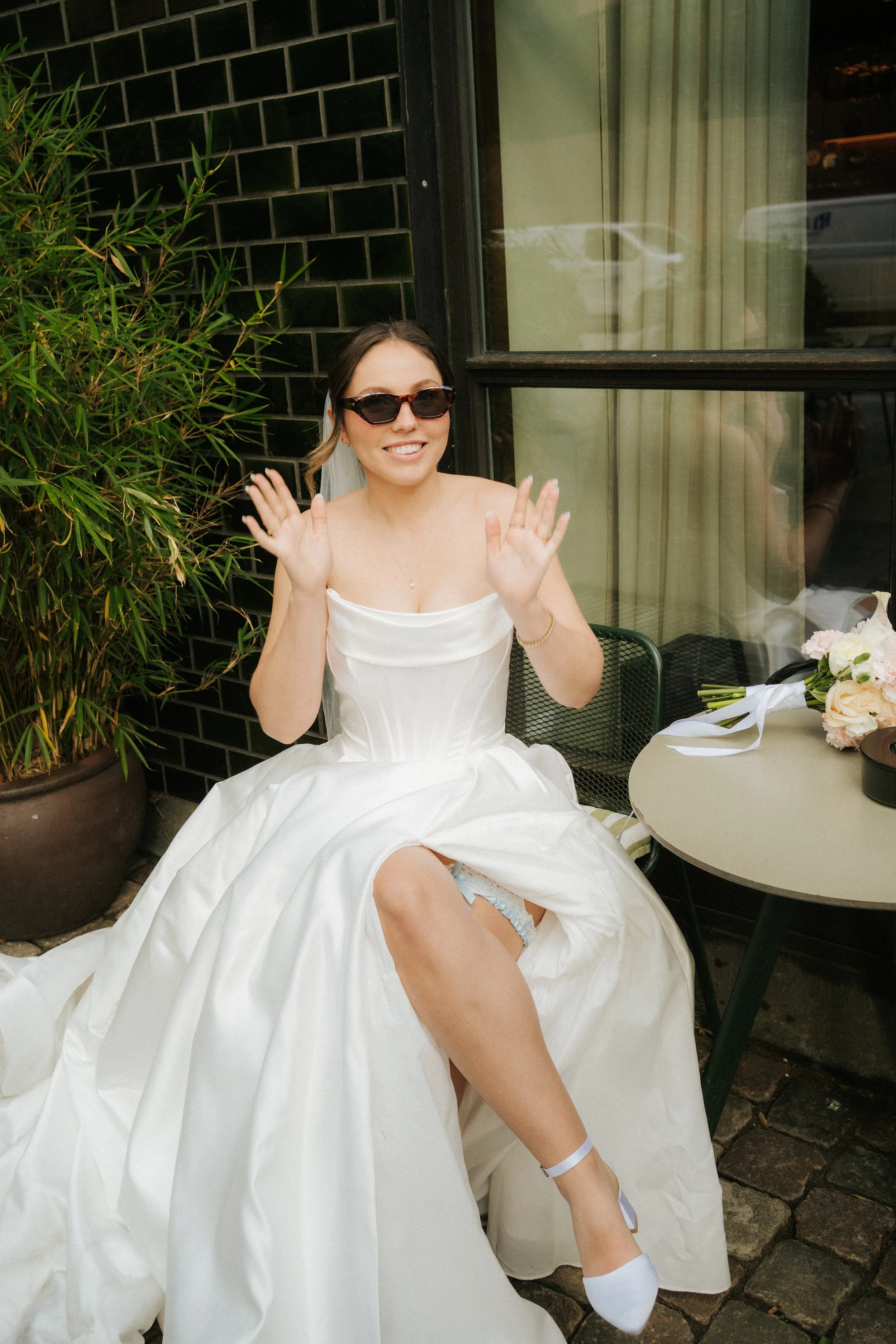 A woman in a wedding dress sitting outside at a table, waving, wearing sunglasses, with a flower bouquet on the table. She is smiling and appears joyful.