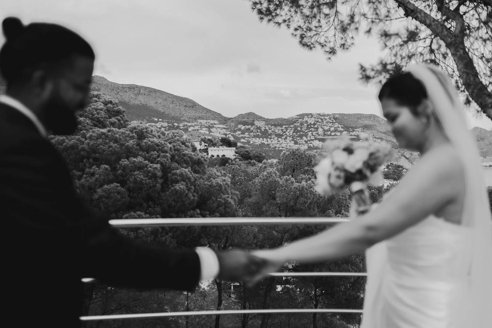 Black and white photograph of a bride and groom holding hands during their wedding ceremony outdoors, with mountains and trees in the background.