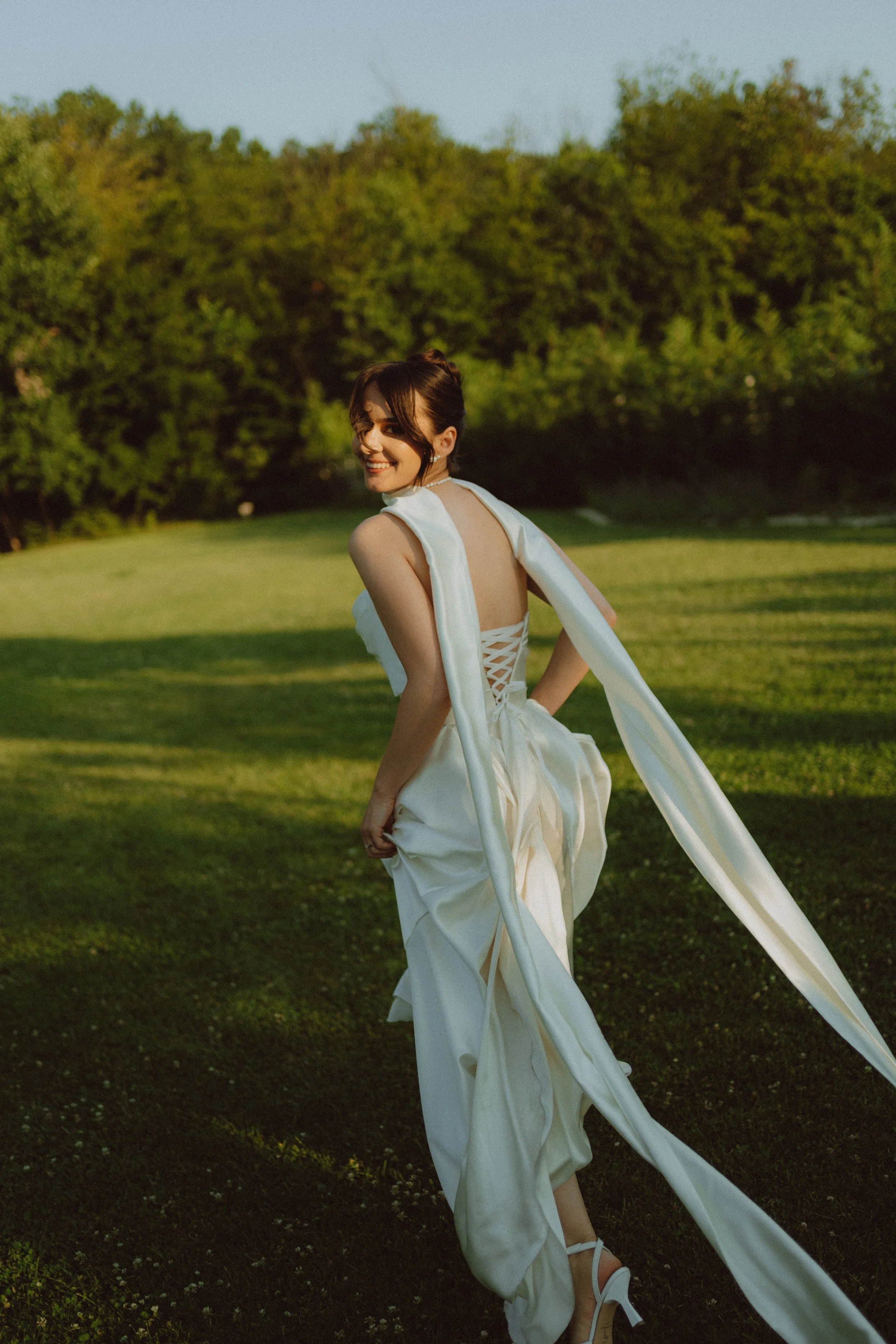 A woman in a white dress standing on grass in a park, smiling, with trees in the background during golden hour.