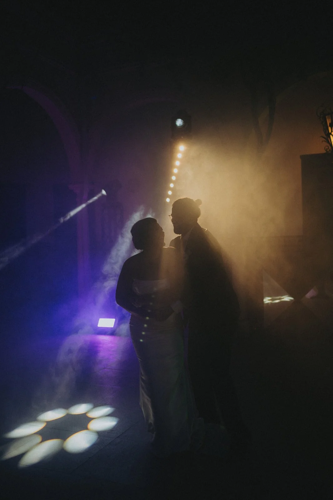 Silhouettes of a man and woman dancing together in a dimly lit room, with colorful lights and fog creating a romantic atmosphere.