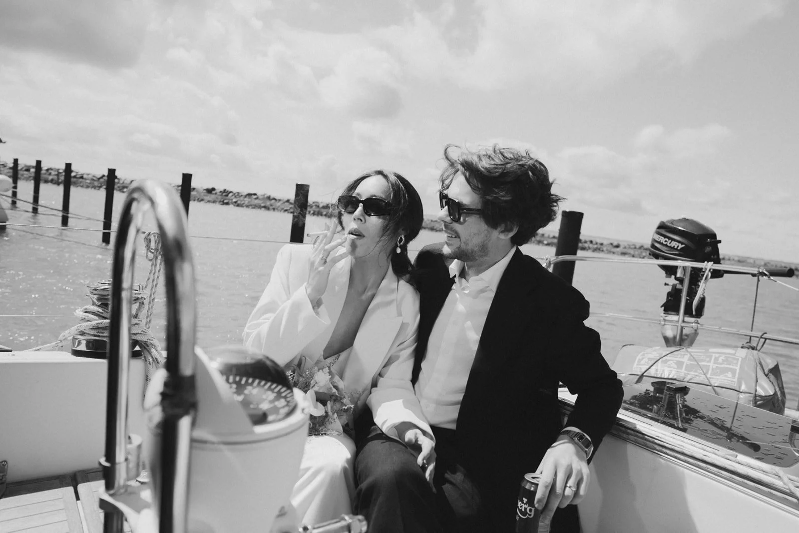 A black and white photo of a man and woman sitting together on a boat. The woman is wearing sunglasses and a white jacket, smoking a cigarette. The man is wearing sunglasses and a dark jacket, holding a drink. The background shows water, a partly clo
