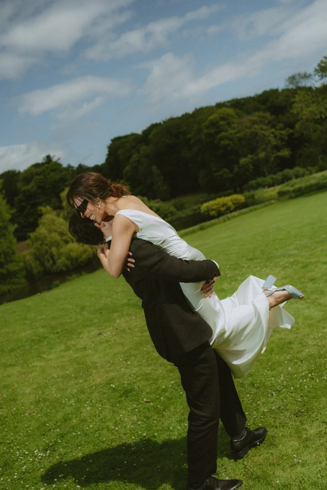 Two women in formal attire embracing outdoors on a grassy field, with trees and a partly cloudy sky in the background.