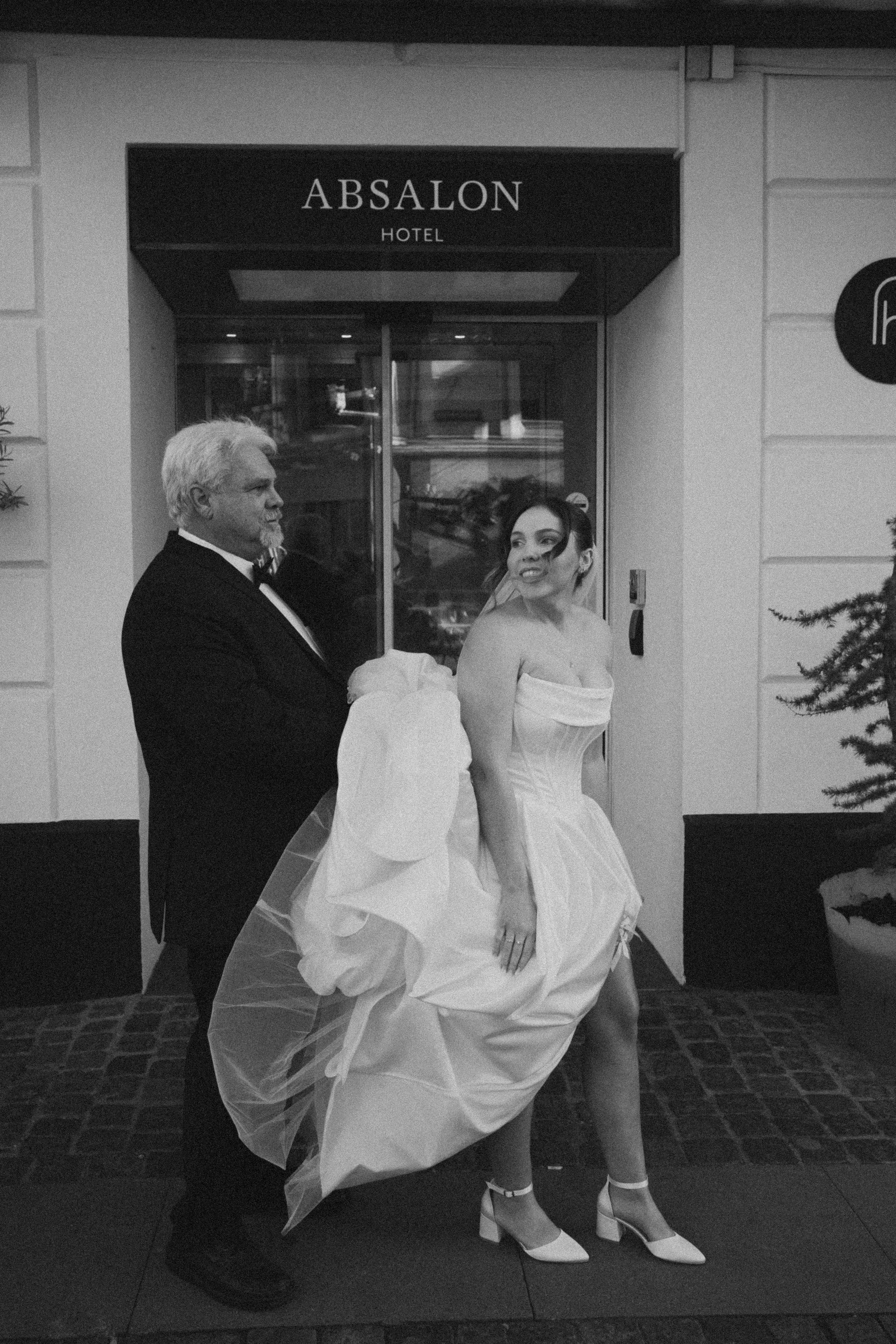 A elderly man in tuxedo helping a young woman dressed in a wedding gown, standing in front of a hotel entrance. The woman is wearing high heels and a strapless wedding dress, and the man appears to be adjusting her dress or veil.