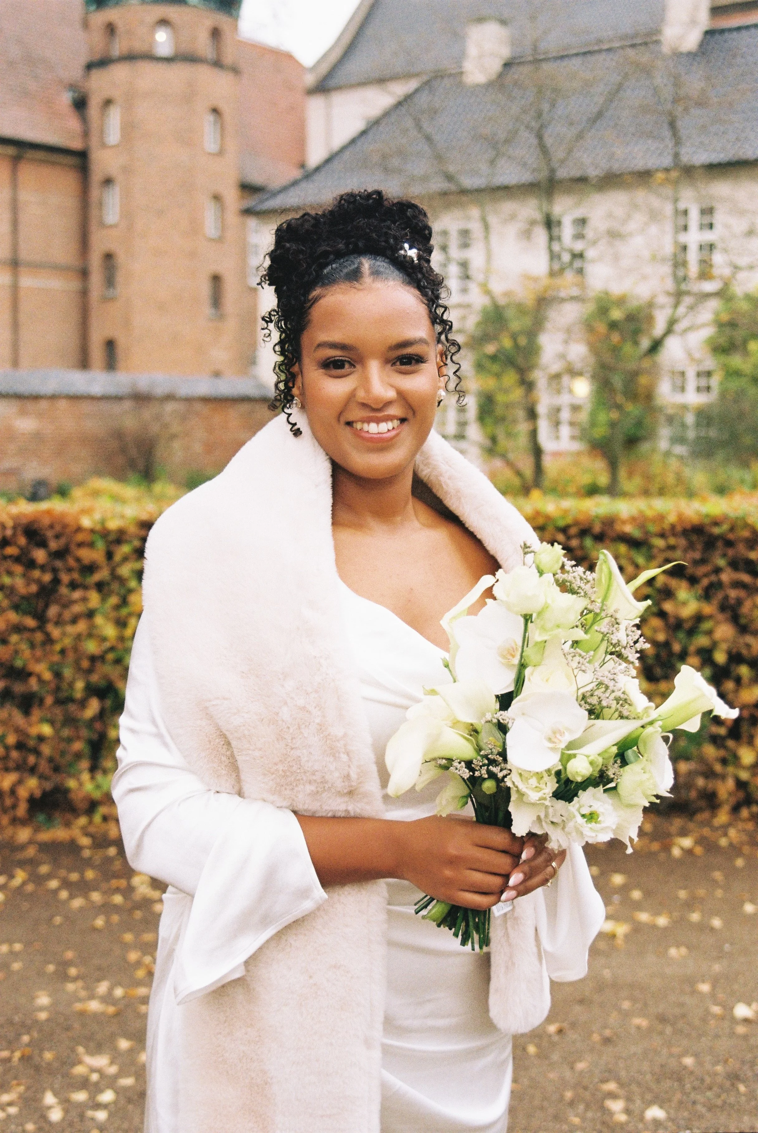 A smiling woman in a white dress and coat holding a bouquet of white flowers outdoors with historic buildings and autumn leaves in the background.