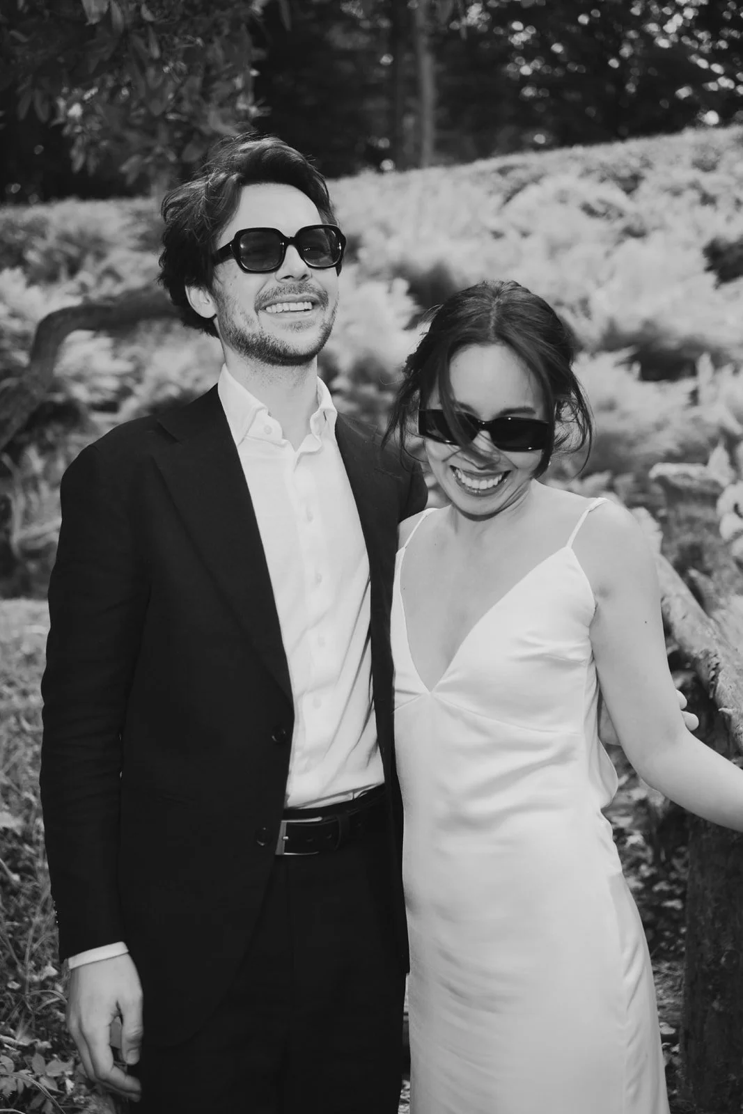 A black and white photo of a man and woman smiling, wearing sunglasses, outdoors with trees and rocks in the background.